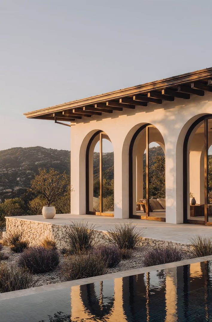 Modern house exterior with white arched columns and large glass doors next to a pool reflecting the architecture, surrounded by plants and hills in the background during sunset.