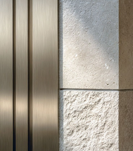 Close-up view of a wall section showing two vertical brushed metal panels on the left and textured stone tiles on the right, with sunlight casting shadows.