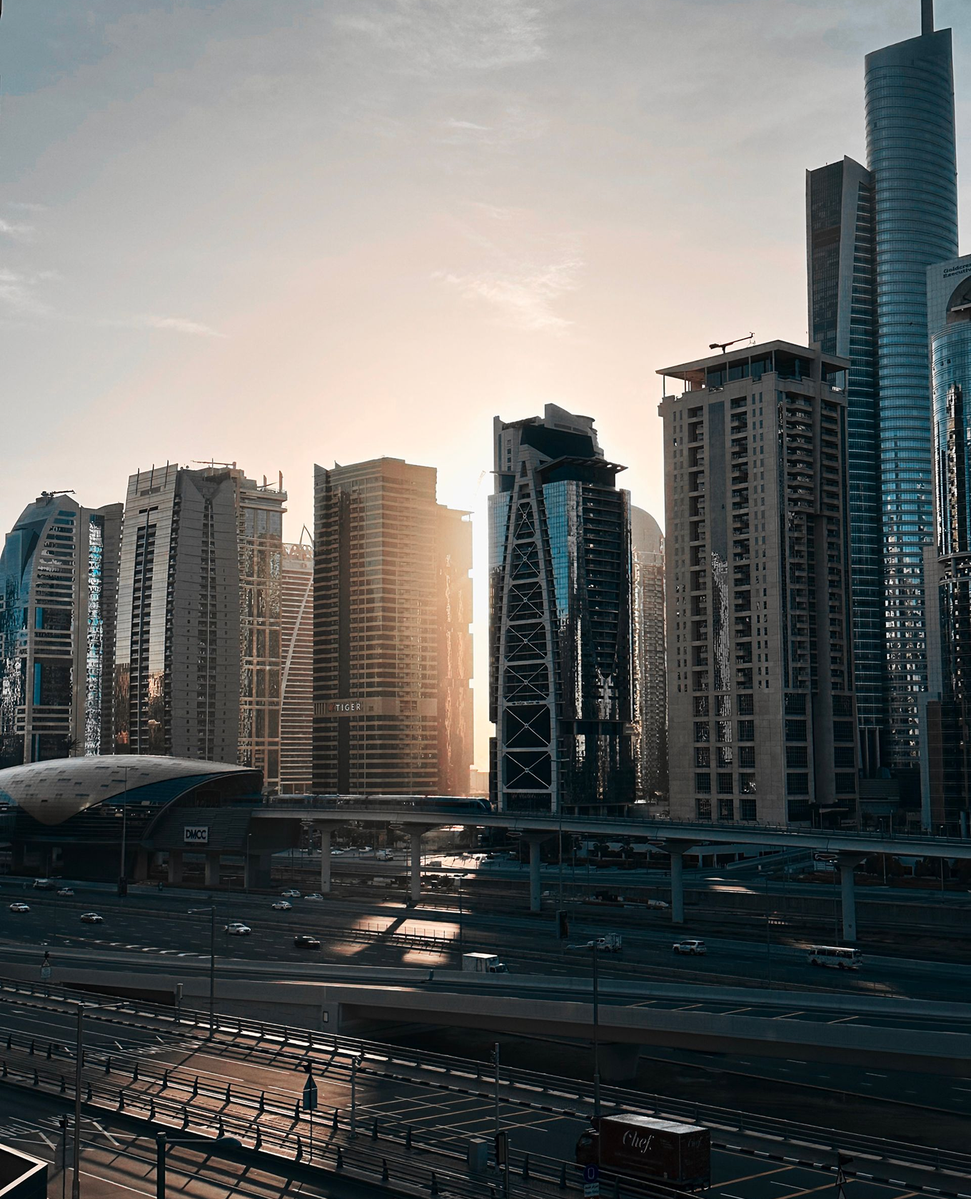 Modern city skyline at sunrise with multiple high-rise buildings reflecting the sun, highways with light traffic, and an elevated metro track with a moving train.