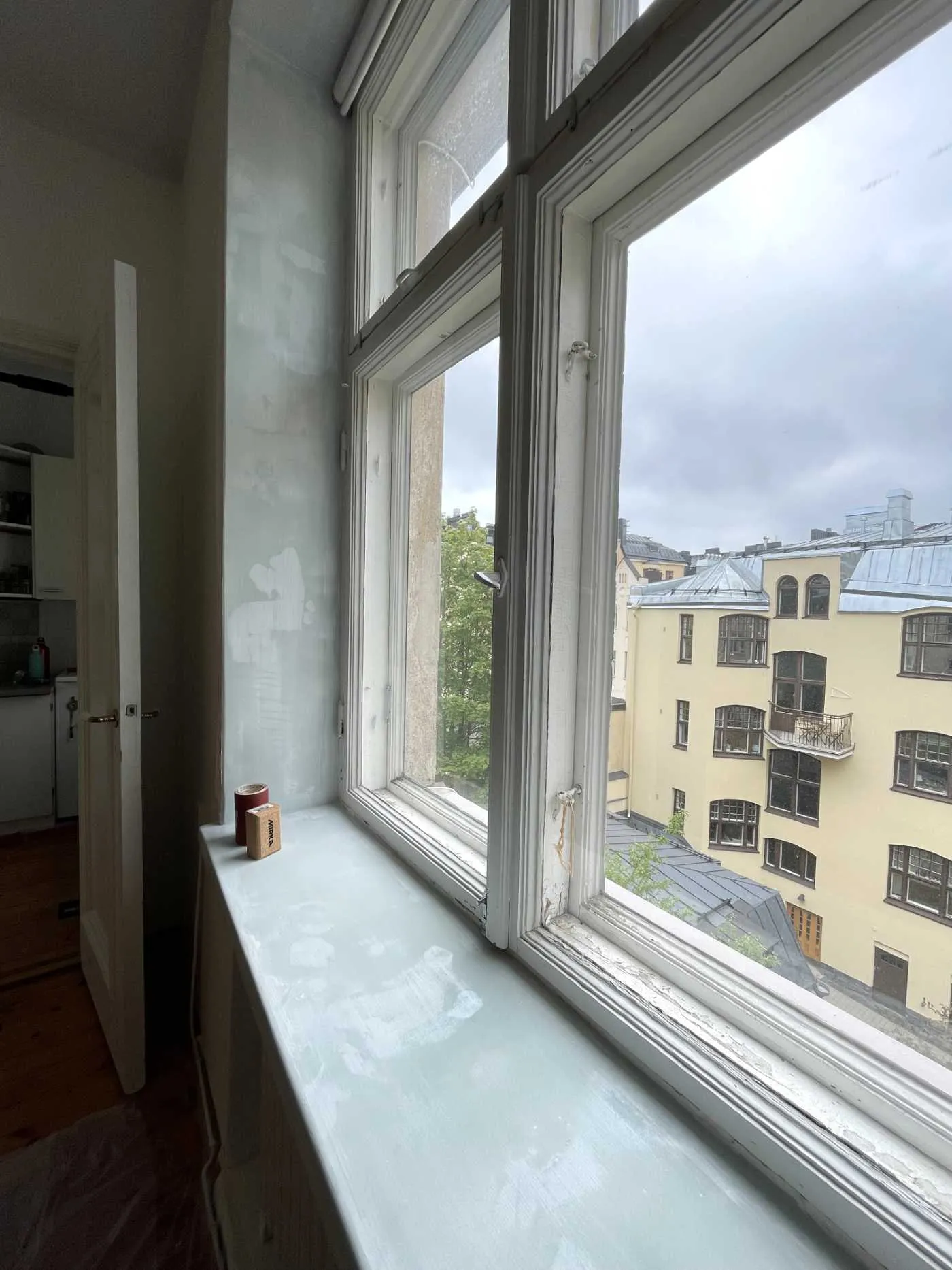 Huoneistoremontin kohteena oleva ikkunanurkka, jossa näkyy osittain maalattu seinä ja ikkunalauta sekä ulkona keltainen rakennus.