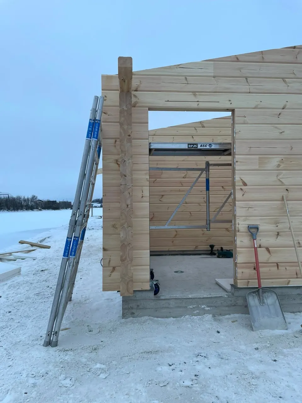 Keskeneräinen puurakennus lumisessa maisemassa, jossa tikkaat nojaavat seinään ja lapio on oven vieressä.