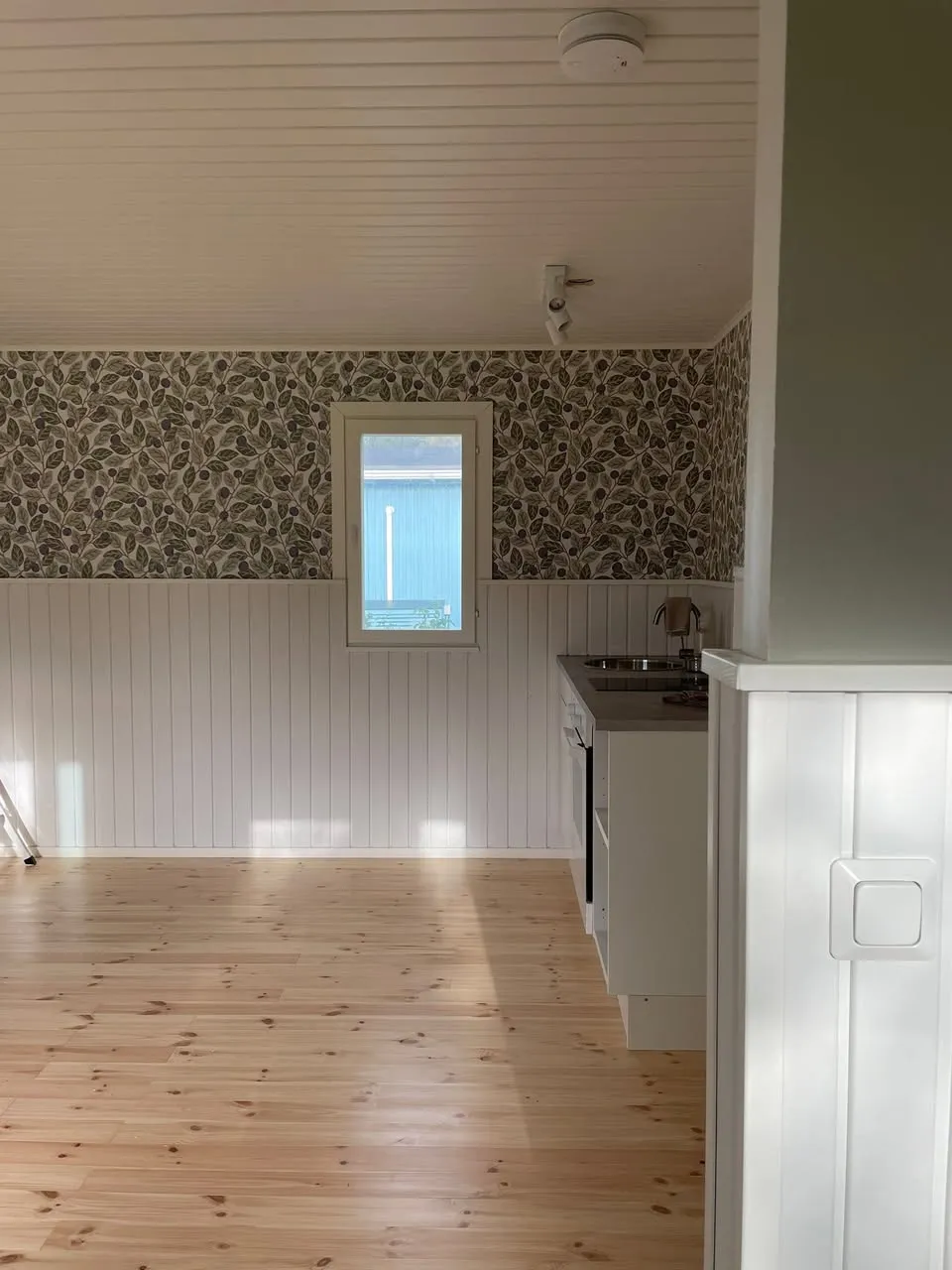 Vastaremontoitu huone, jossa on vaalea puulattia, valkoiset paneeliseinät ja kuviollinen tapetti.