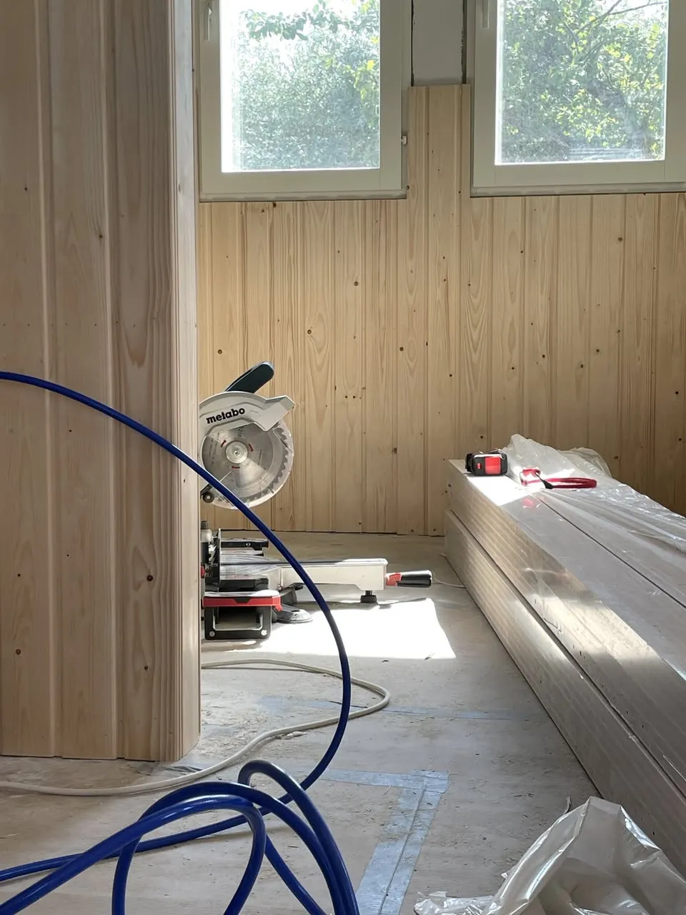 Huoneistoremontin keskellä näkyy puupaneeliseinä, kaksi ikkunaa, katkaisusaha ja pinottuja rakennusmateriaaleja lattialla.