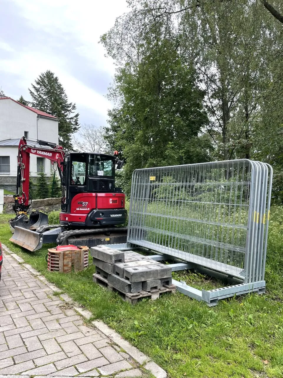 Punainen kaivinkone ja remonttiin liittyviä rakennustarvikkeita, kuten betonilaattoja ja metalliaitoja, on asetettu nurmikolle kivetyn polun viereen.