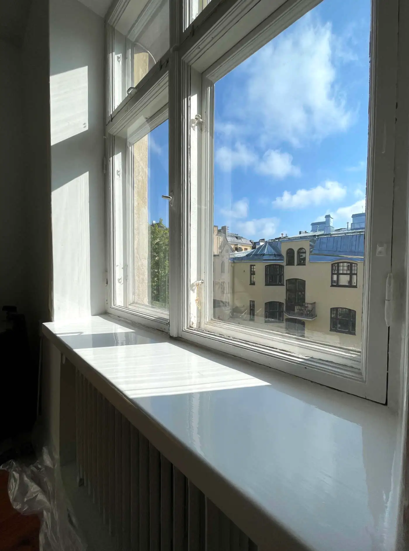 Aurinko paistaa valkoisesta ikkunasta ja ikkunalaudasta, joista avautuu näkymä kaupungin rakennuksiin sinisen taivaan alla huoneistoremontin jälkeen.