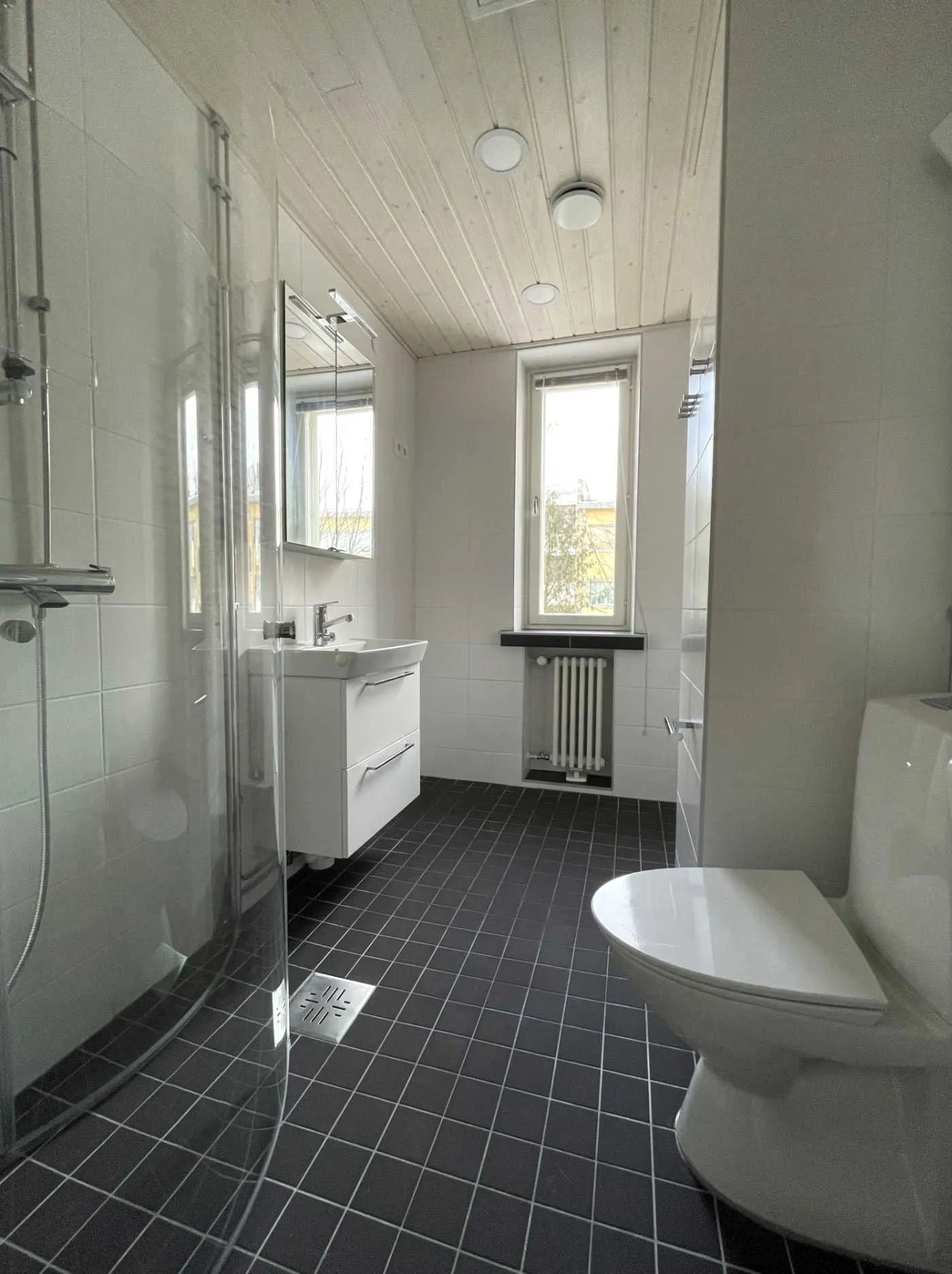 Kylpyhuone, jossa on valkoiset seinälaatat, tummat lattialaatat, suihku, pesuallas ja wc, on tyypillinen huoneistoremontin kohde.