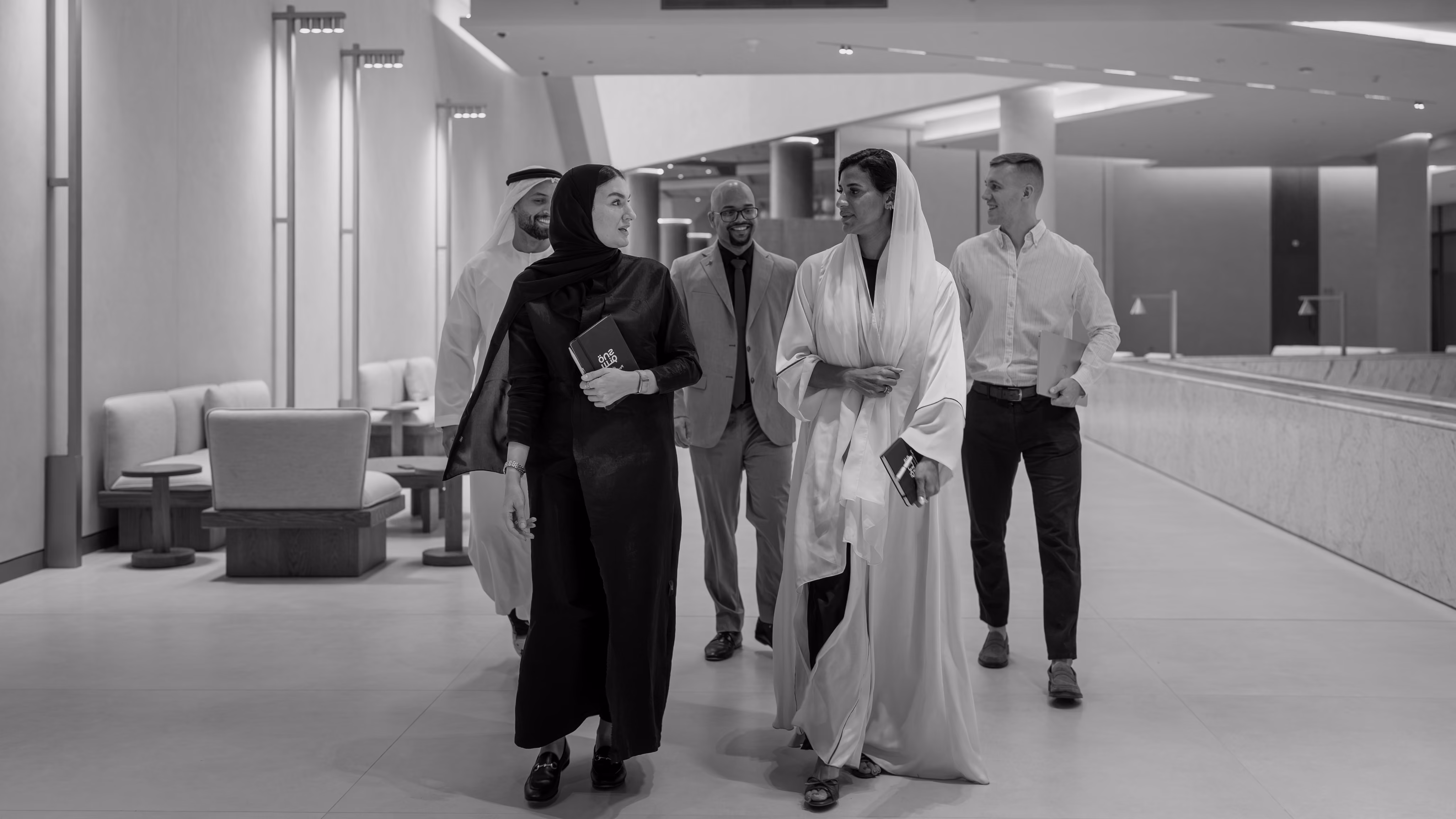 A black and white image of a group of men and woman walking in a hallway, each wearing different cultural attire.
