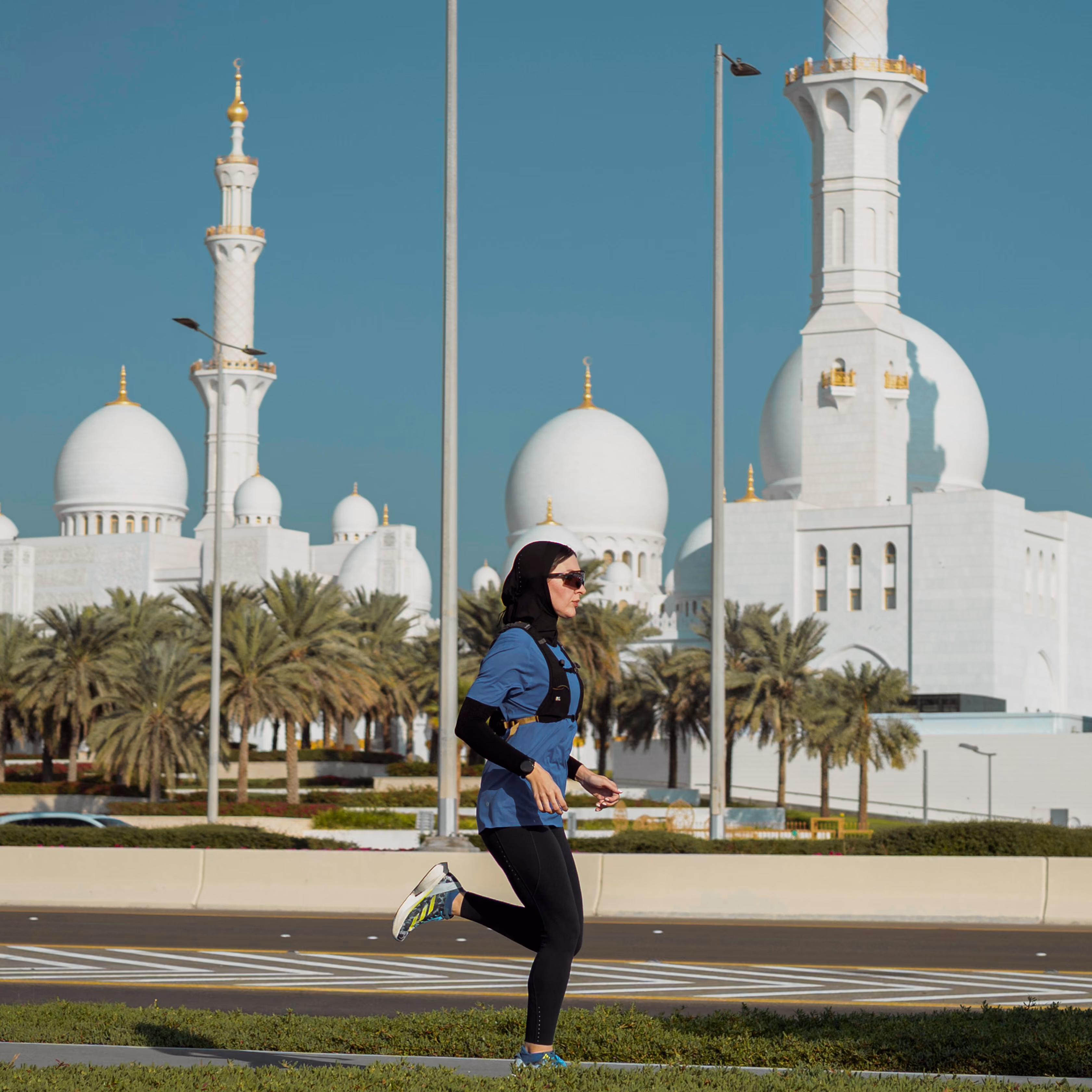A woman wearing running attire, running on a sunny day past a fully white Mosque.