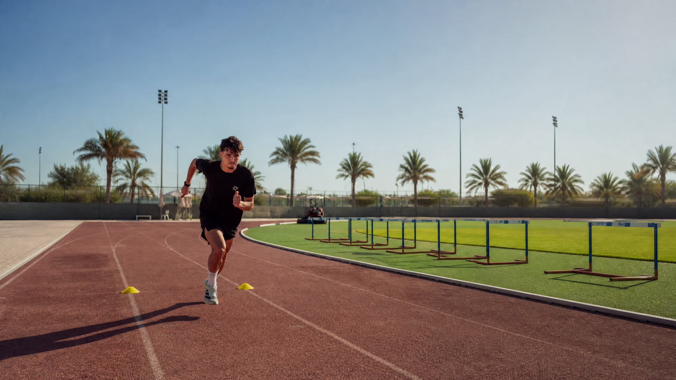 Athlete sprinting on a palm-lined running track beside a row of hurdles