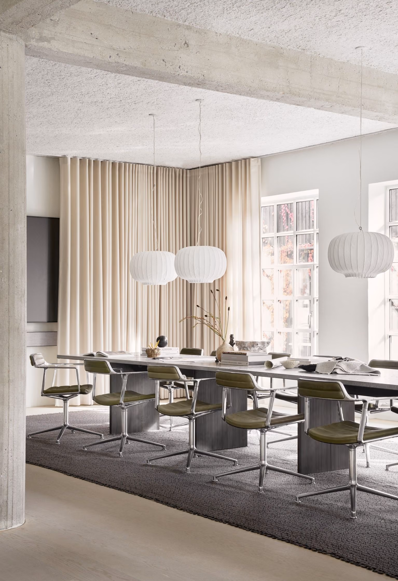 Modern dining area with a long black table surrounded by green swivel chairs with chrome legs on a dark rug, white textured pendant lamps hanging above, and large windows with beige curtains allowing natural light.