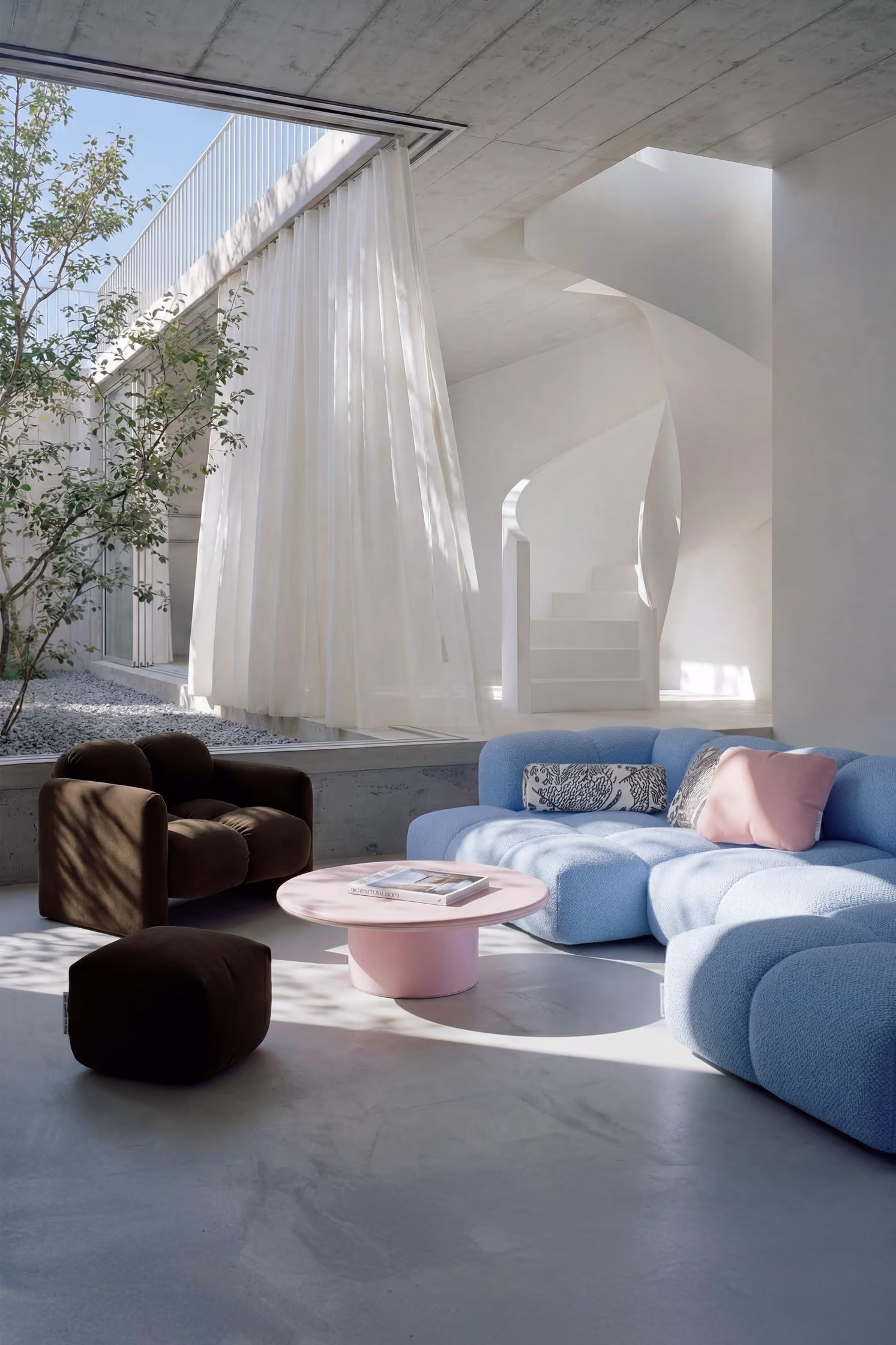 Modern living room with a light blue sectional sofa, pink and patterned cushions, a dark brown armchair, matching ottoman, and a round pink coffee table with a book on it, bathed in natural light from large windows with sheer white curtains next to a minimalist white spiral staircase and indoor tree.