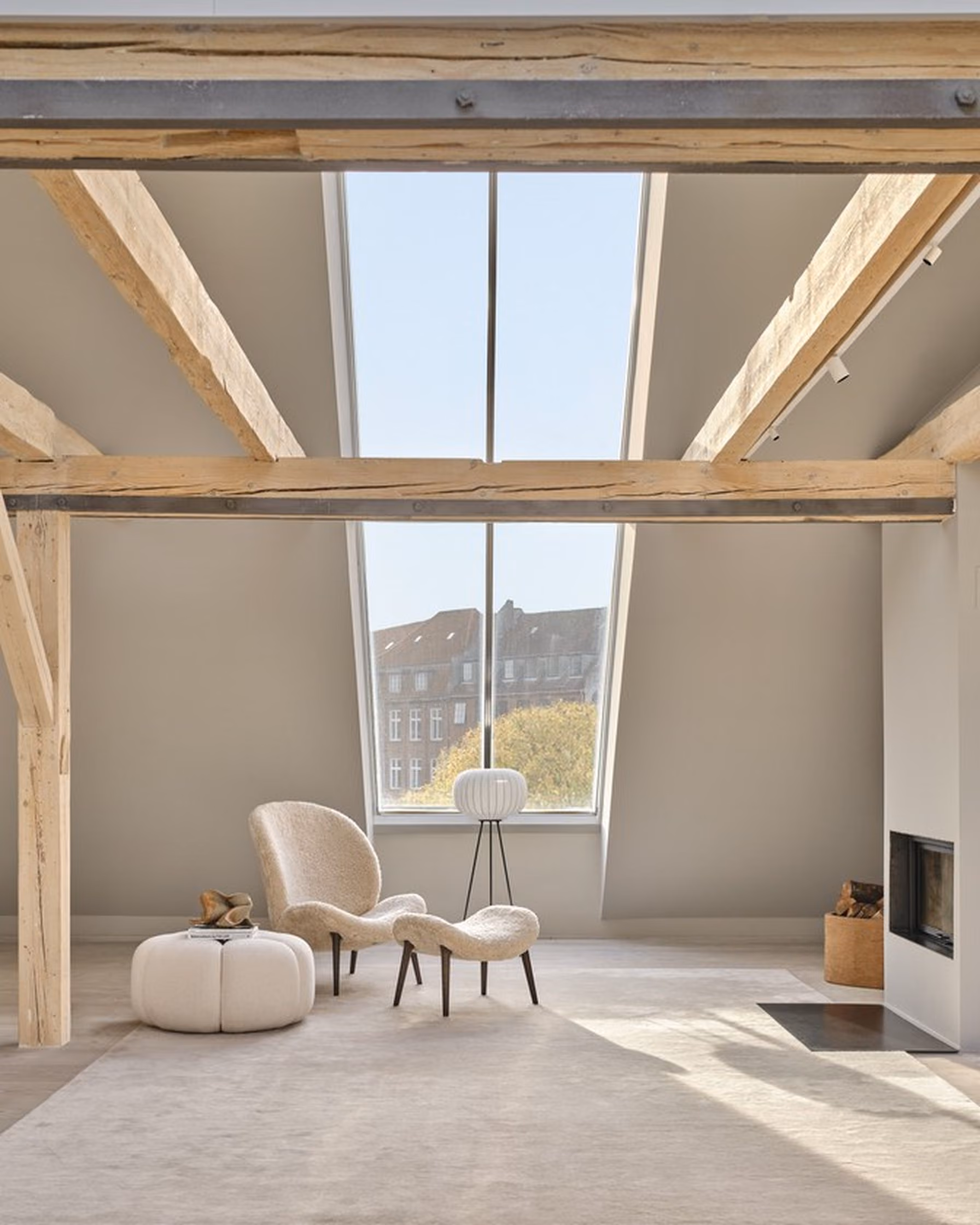 Minimalist attic room with exposed wooden beams, a large central skylight window, a plush beige armchair with matching footstool, a round white ottoman, a tripod floor lamp, and a modern fireplace on the right.