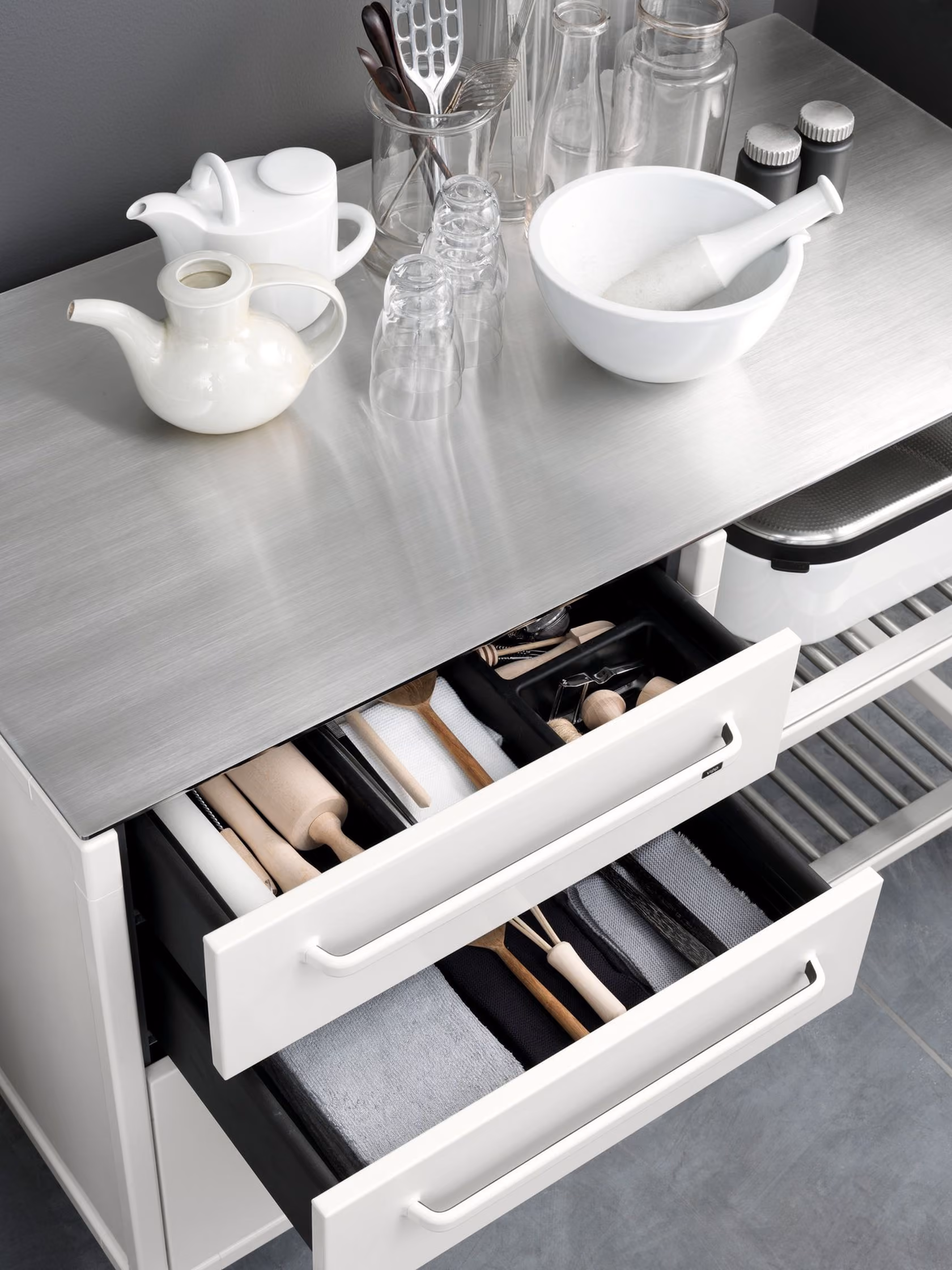 Modern kitchen stainless steel countertop with white ceramic teapots, glass jars, a white mixing bowl with a pestle, small glasses, and kitchen utensils; below, two open drawers containing rolling pins, measuring tools, wooden spoons, a whisk, and folded kitchen towels.