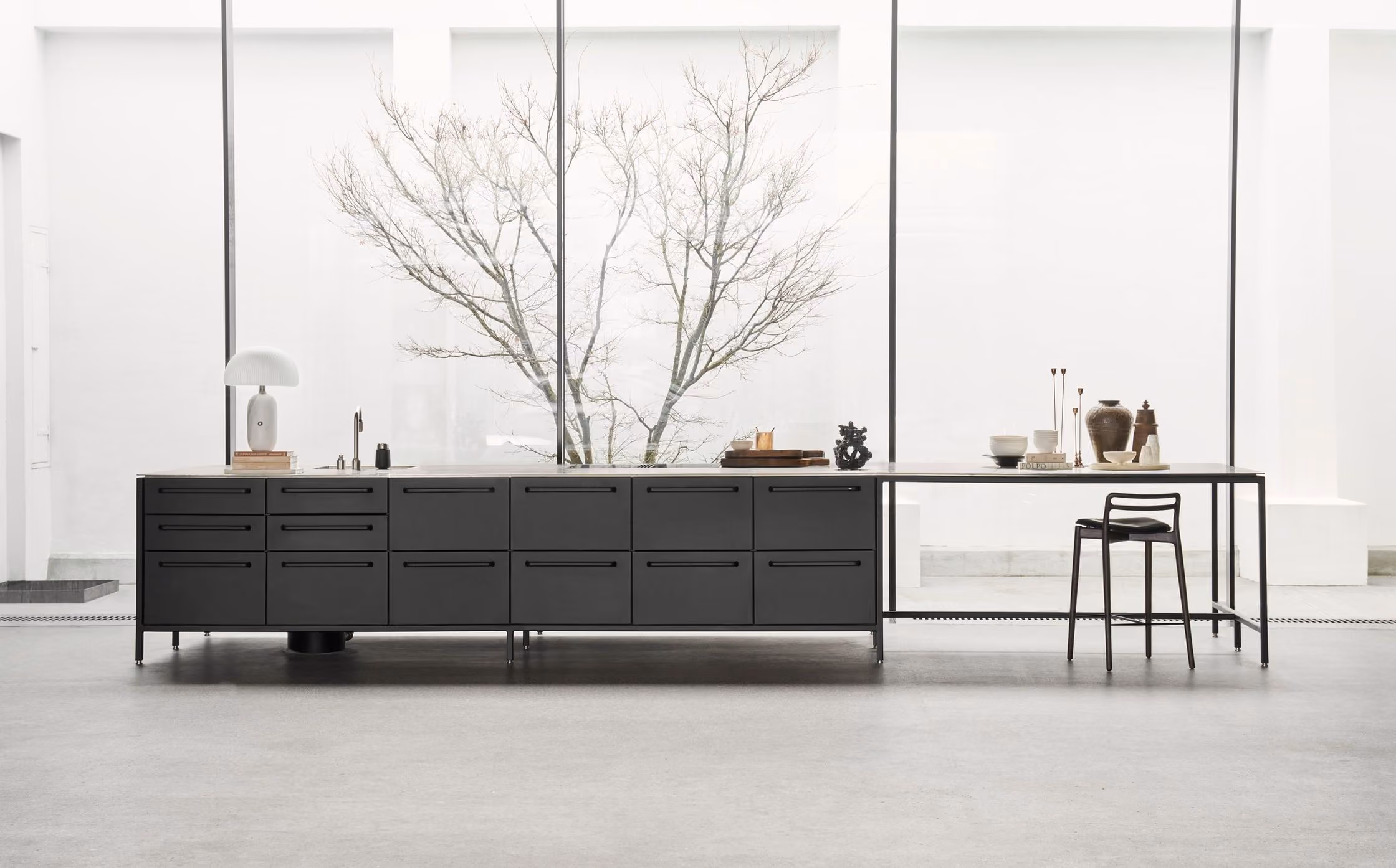 Minimalist modern kitchen island with black drawers and a slim table extension, set against large floor-to-ceiling windows showing a bare tree outside.