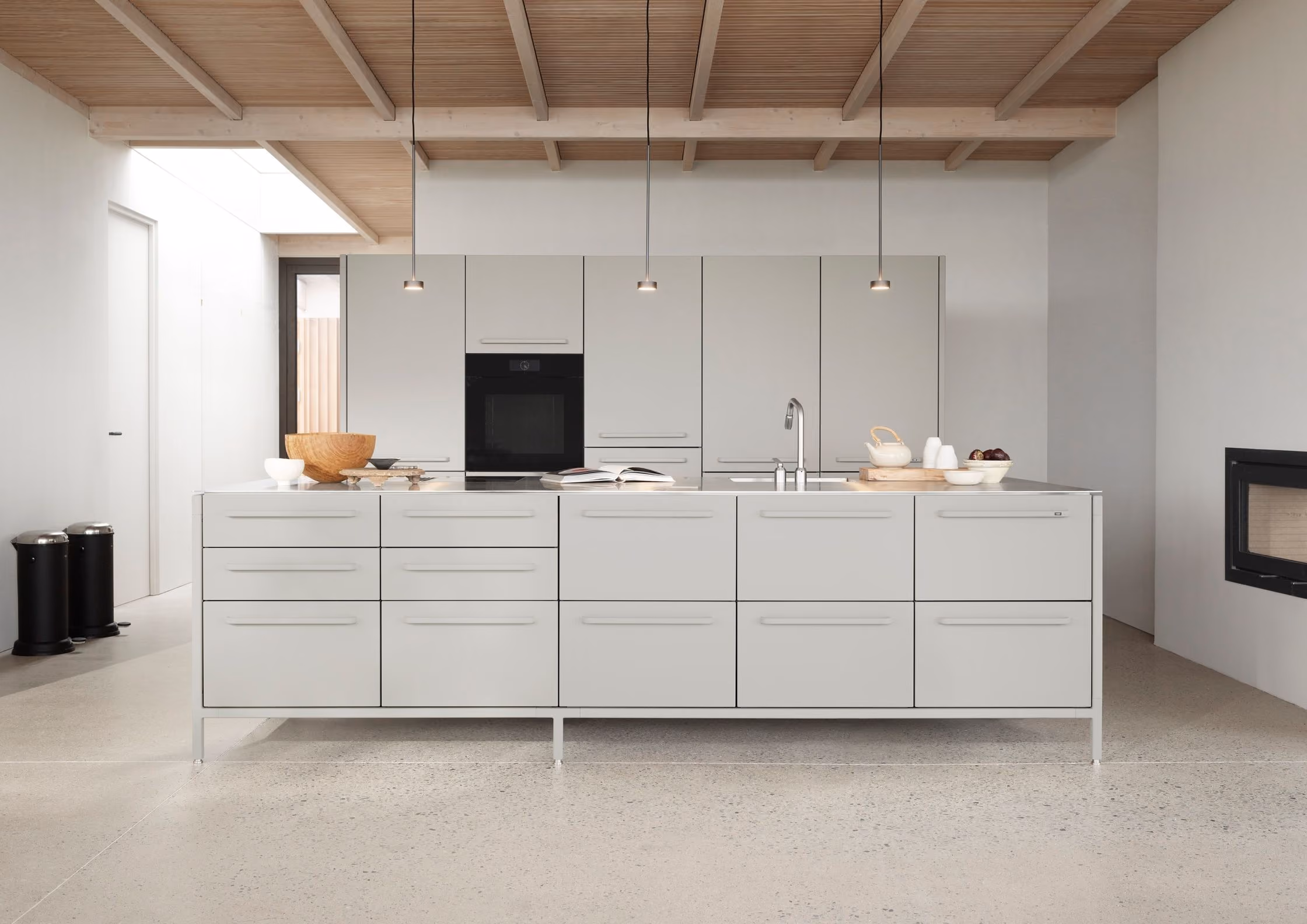 Minimalist modern kitchen with light grey cabinetry, central island featuring a sink, and three pendant lights against a wooden ceiling.