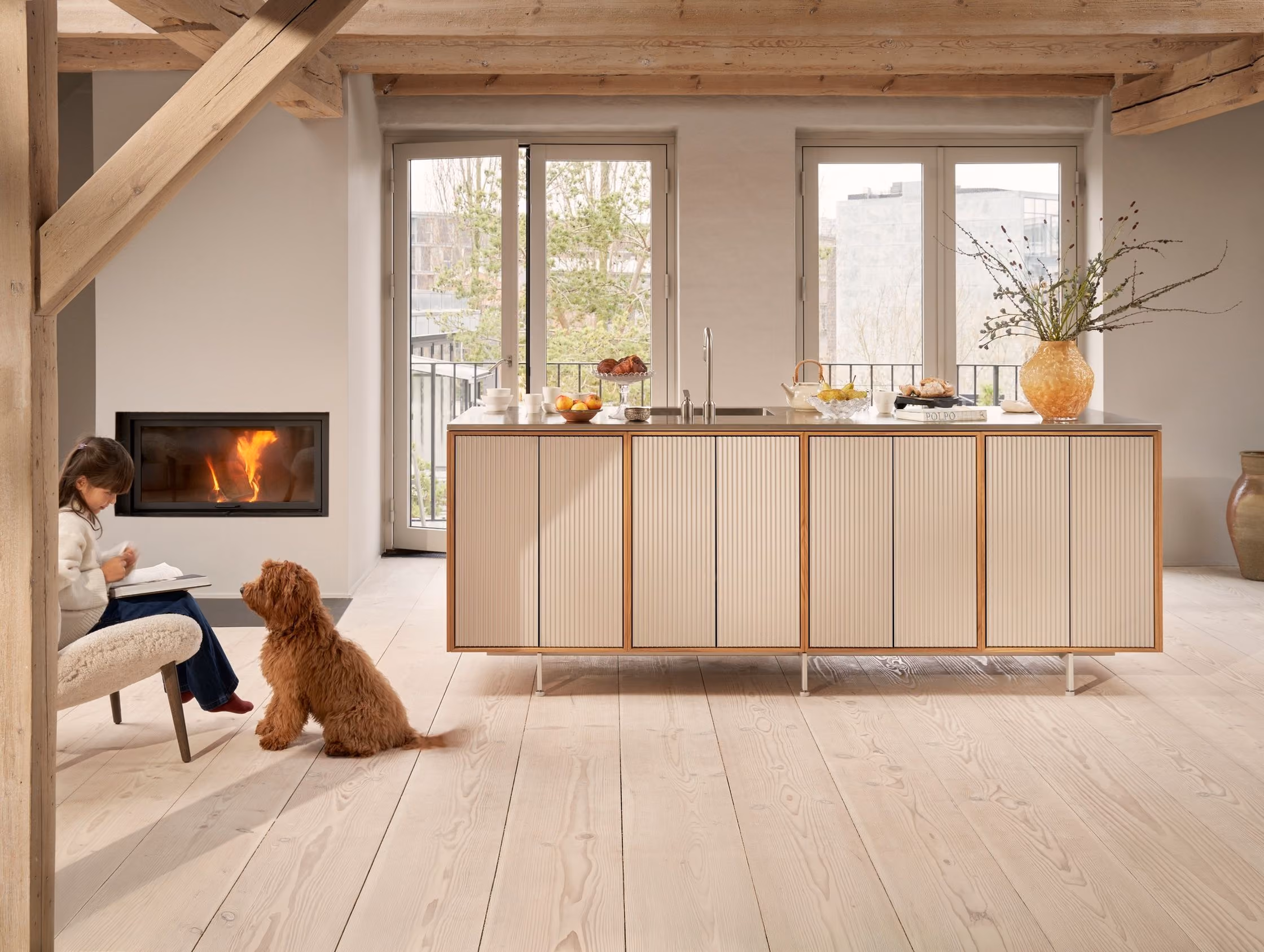 Modern kitchen with light wooden floor and ceiling beams, a large central island with vertical panel cabinets, a vase with branches, fruit bowls, and a seated child beside a brown fluffy dog by a fireplace with a fire inside.