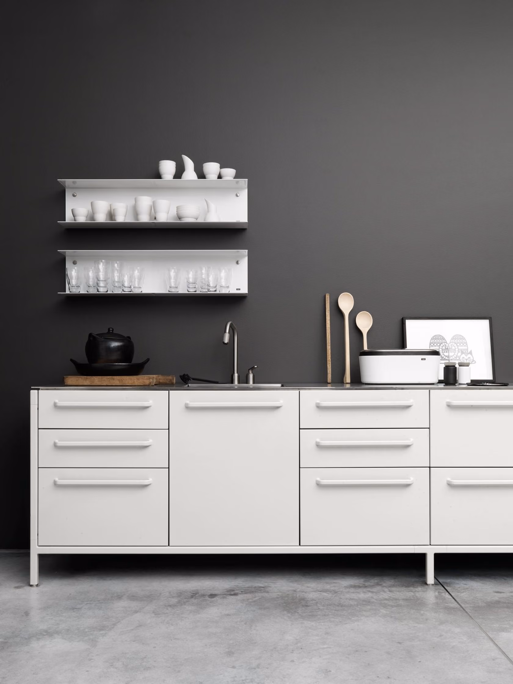 Minimalist white kitchen cabinet with metal handles against dark gray wall, featuring built-in sink, black kettle on wooden cutting board, two white shelves with white cups and glassware, wooden spoons, a white container, and framed monochrome artwork.