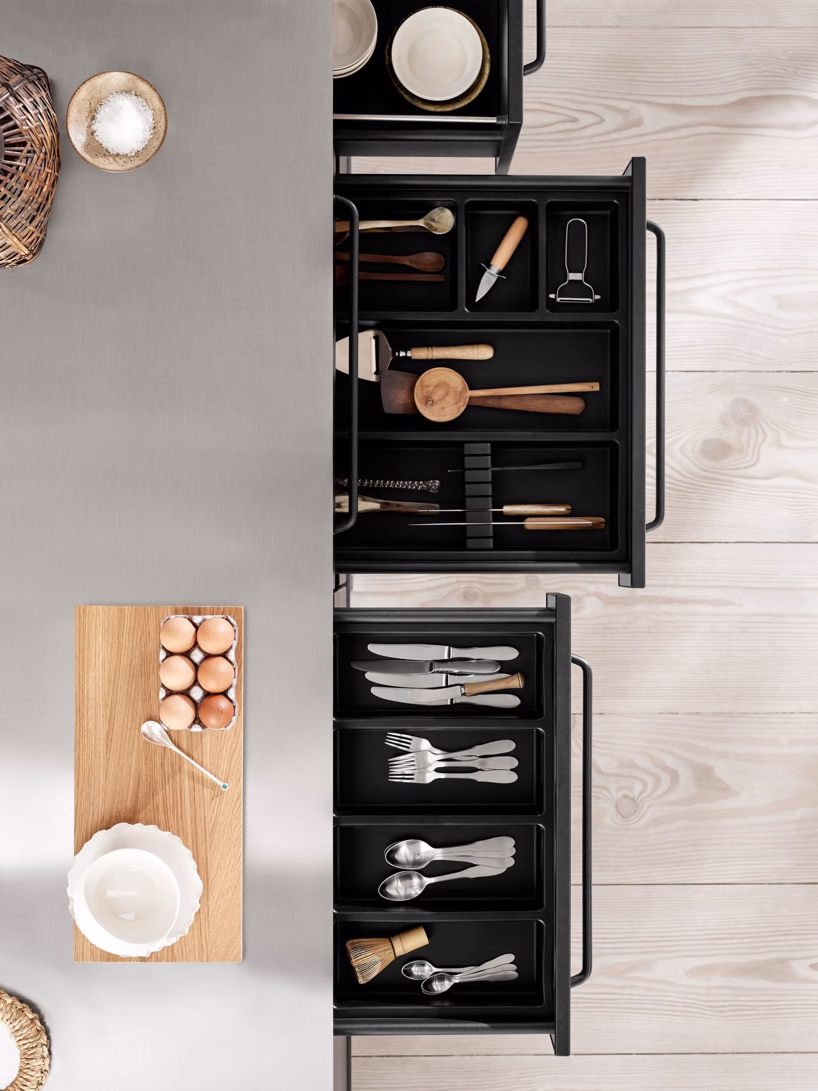 Open kitchen drawers showing organized utensils including knives, forks, spoons, wooden spoons, a vegetable peeler, a small knife, a whisk, and a small brush, alongside a wooden cutting board with a carton of eggs and a white bowl on a light wood floor.