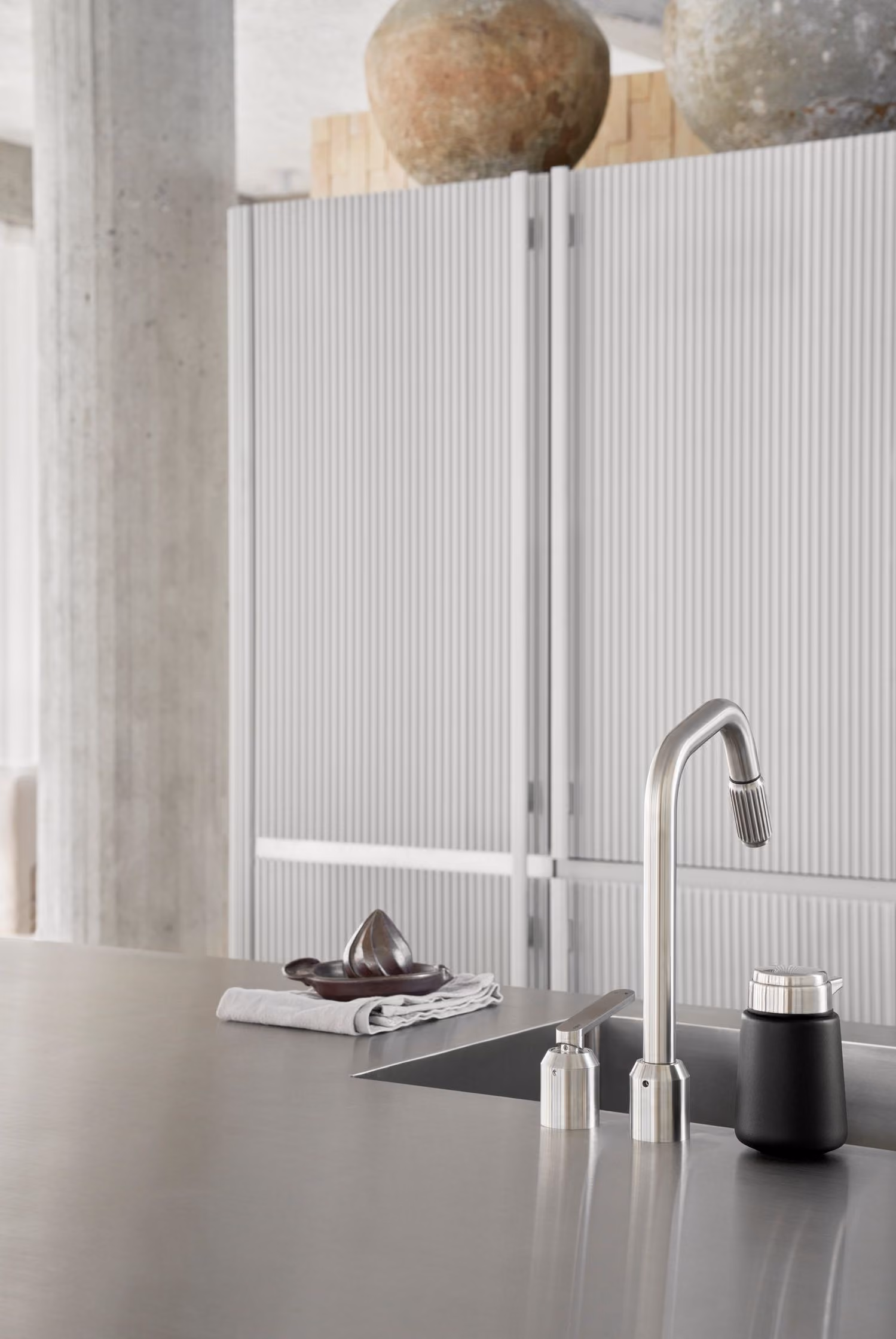 Modern kitchen sink area with a stainless steel faucet, soap dispenser, and a black container on a smooth gray countertop, with textured white cabinets and rustic pottery in the background.
