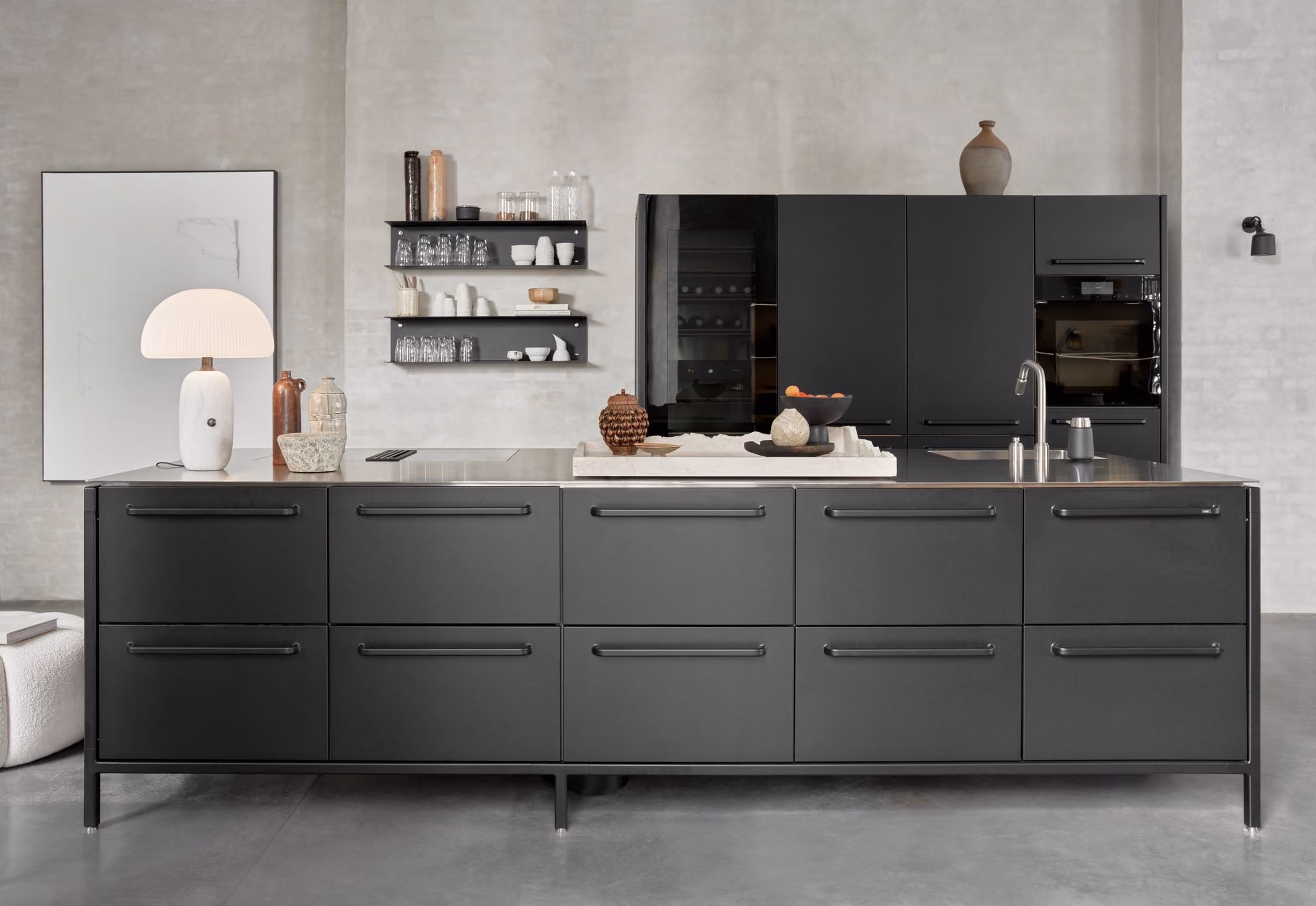 Modern kitchen with minimalist design featuring a large gray island with drawers and a stainless steel countertop including a sink and faucet. On the island are decorative items like a bowl of oranges and textured objects. Behind the island, there is a wall with black built-in cabinets, a built-in oven, and an open wine fridge. Three black floating shelves hold clear glassware and white dishware. On the left side of the image, a white lamp and ceramic vases sit on the counter, next to a large framed picture leaning against the wall. The kitchen has a polished concrete floor and light gray walls.