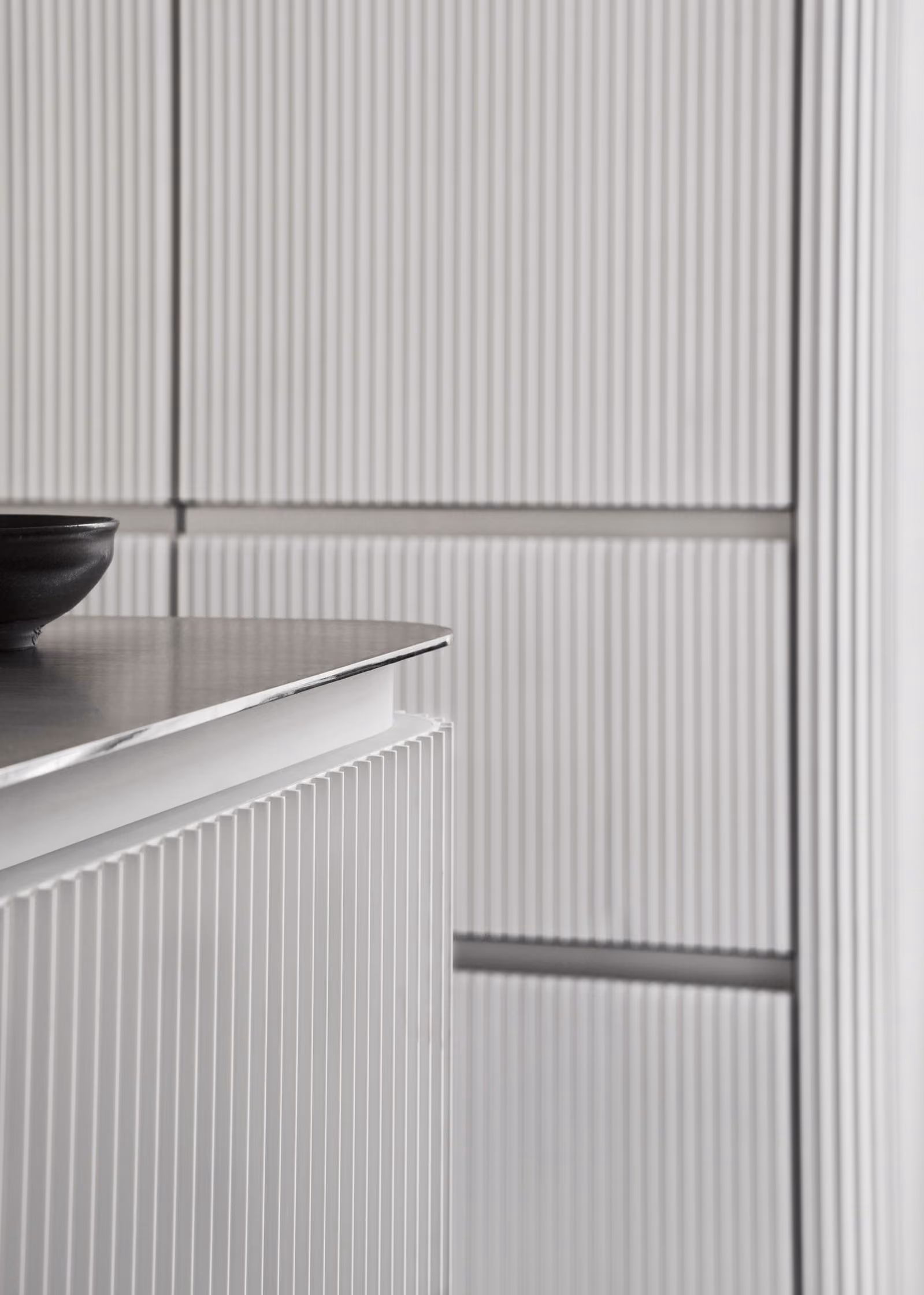 Close-up of a modern kitchen with vertically grooved white cabinetry and a sleek metallic countertop with a black bowl on it.
