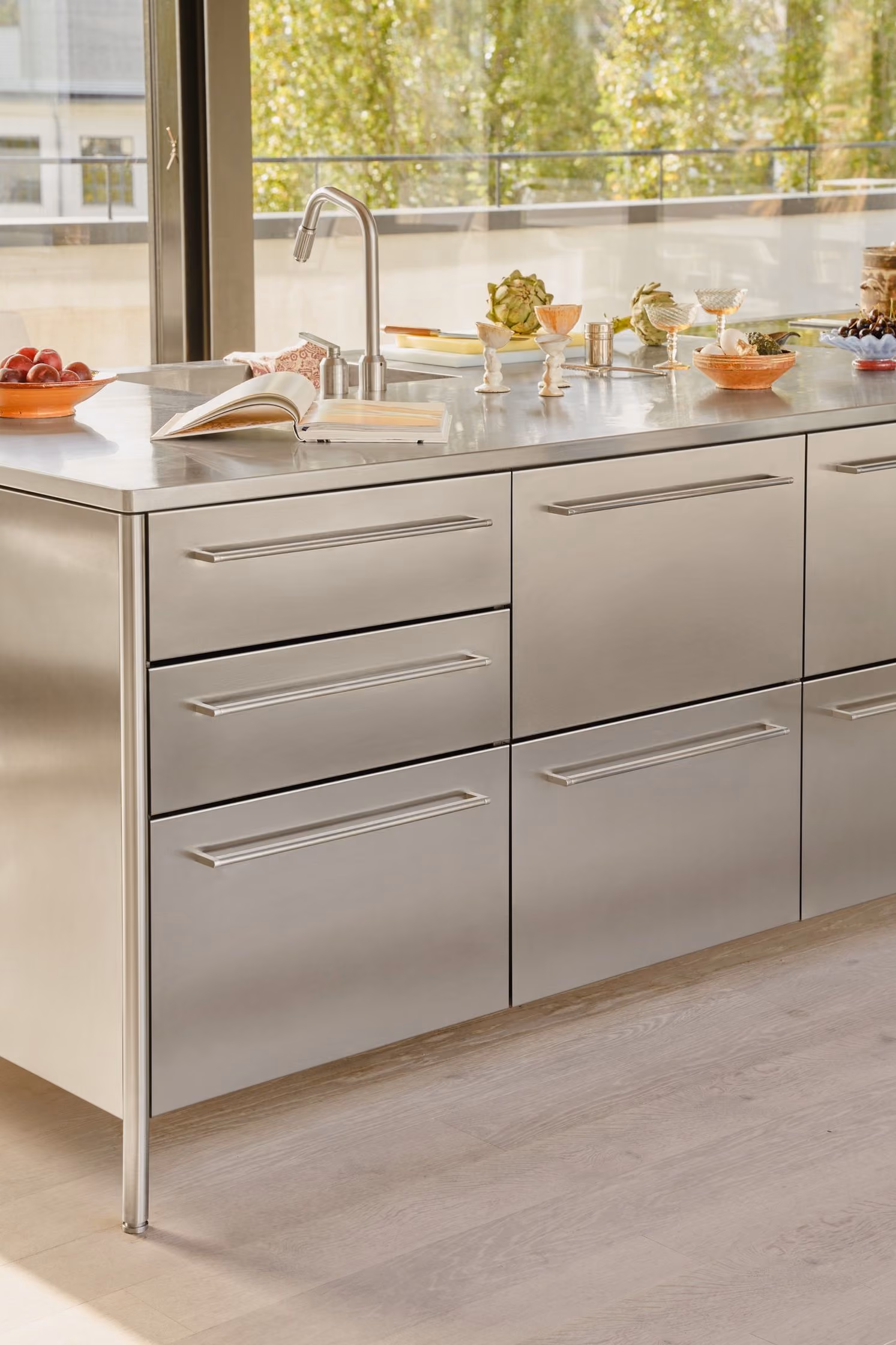 Modern kitchen island with stainless steel cabinets and drawers under a countertop holding a faucet, open book, and various bowls and glassware containing fruits and decorative items near a large window overlooking greenery.