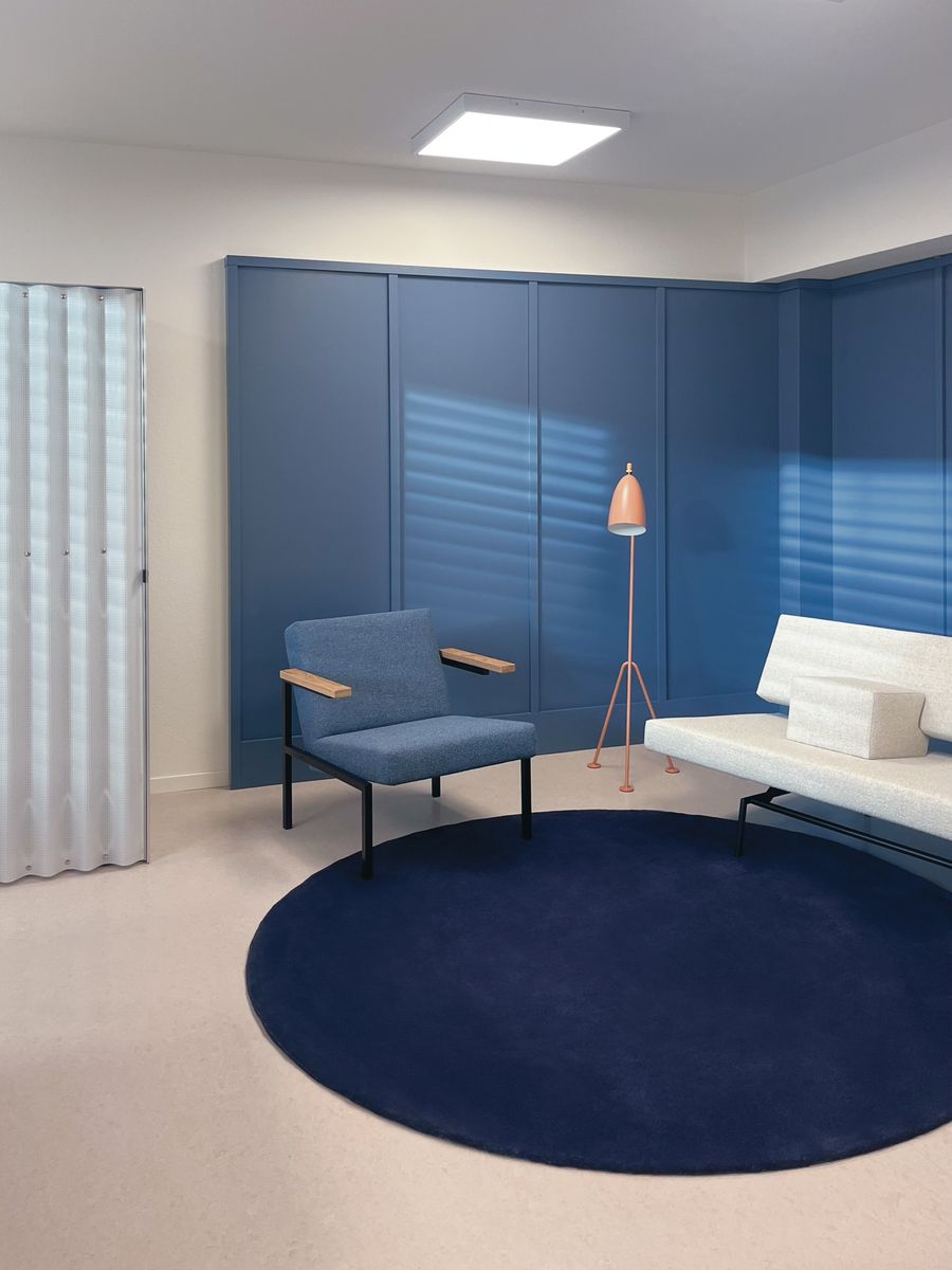 Minimalist modern room with blue paneled walls, a blue armchair with wooden armrests, a white sofa with a cushion, a tall peach-colored floor lamp, and a large navy circular rug on a light floor.