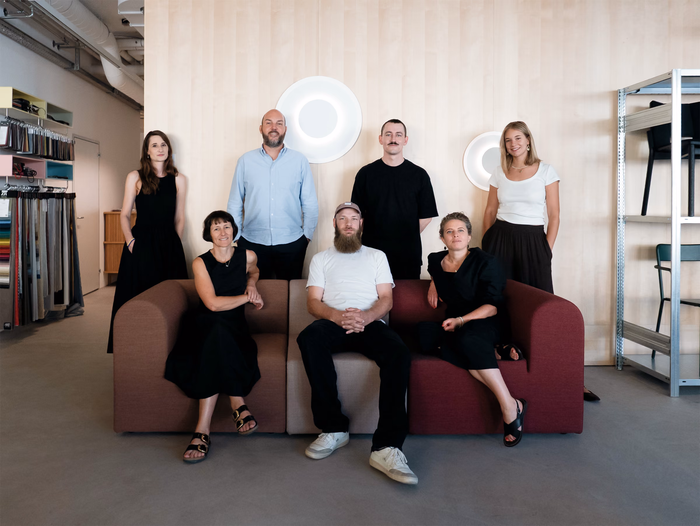 Group portrait of seven adults posing in a modern, minimalistic room with light wood walls and industrial elements; three people are seated on a burgundy and brown sofa, and four stand behind them.