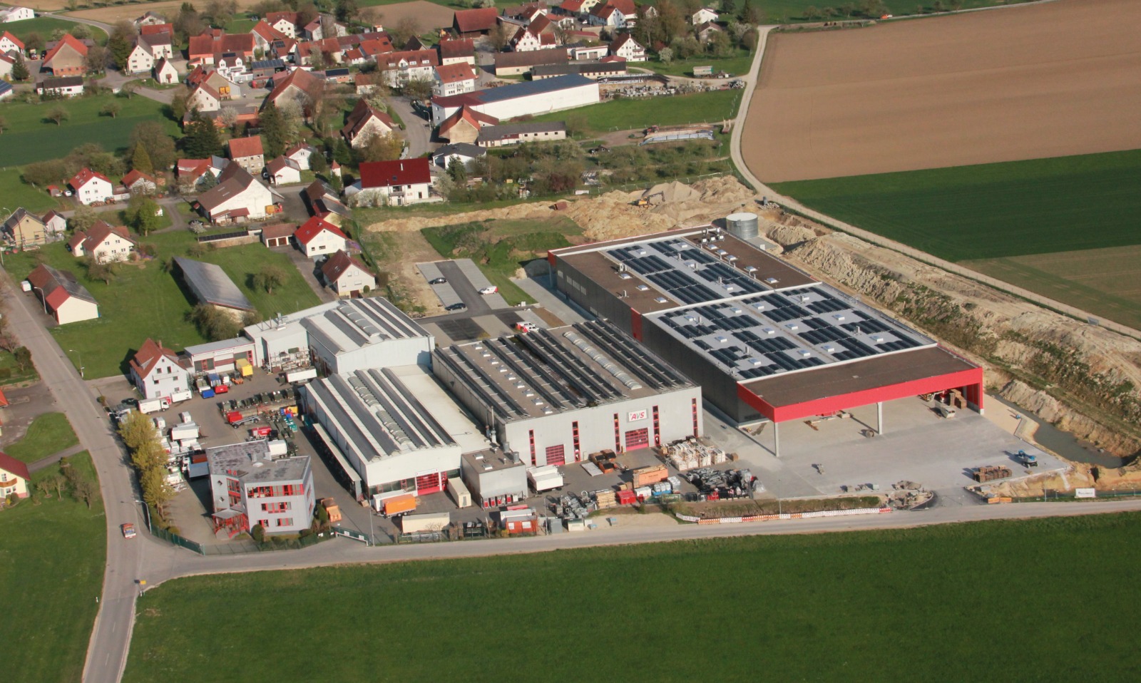 Luftaufnahme des AVS-Werksgeländes mit Fabrikhallen und Solardach, umgeben von einem Dorf und landwirtschaftlichen Feldern