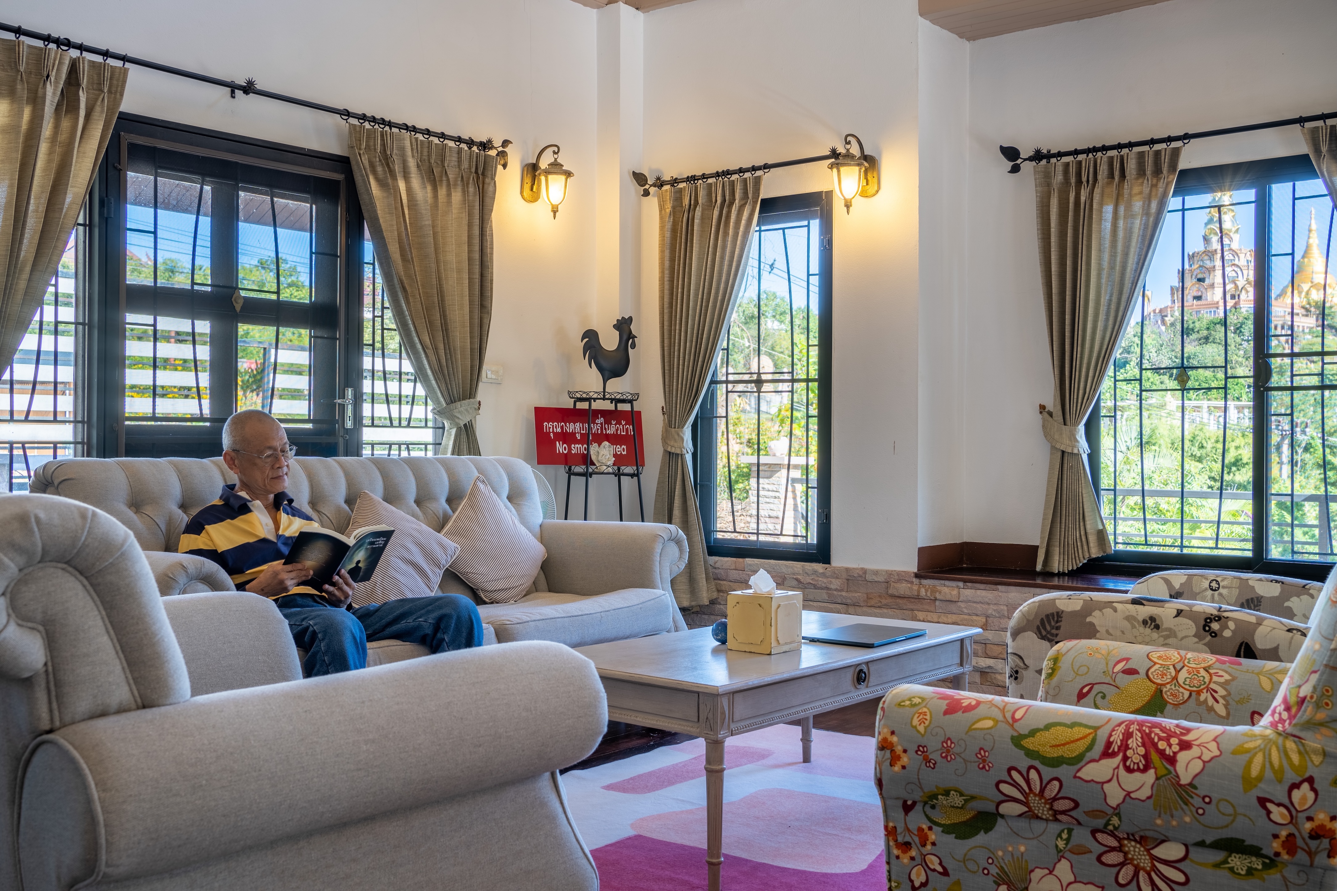 Elderly man reading on a beige sofa in a bright living room with large windows and floral chairs of Verryn Khao Kho's house.