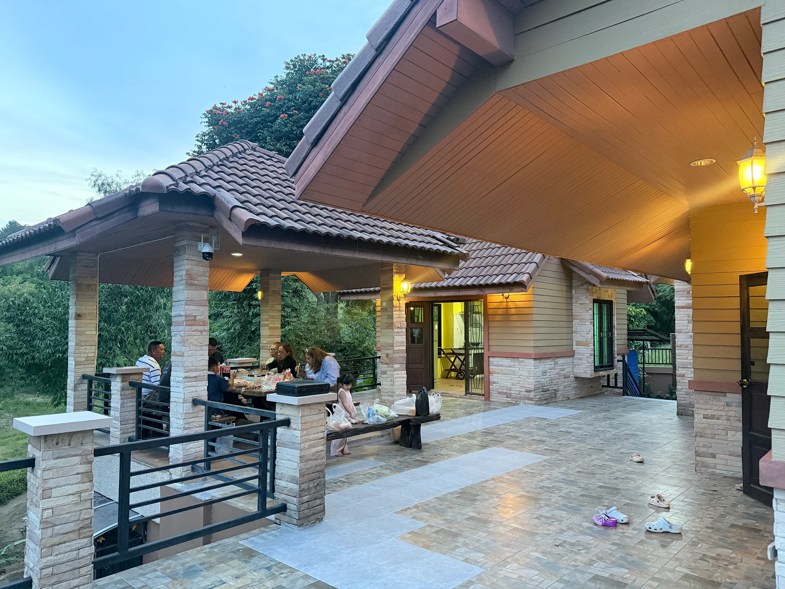 Family gathered under a tiled roof pavilion beside a house at dusk with shoes scattered on the tiled floor.