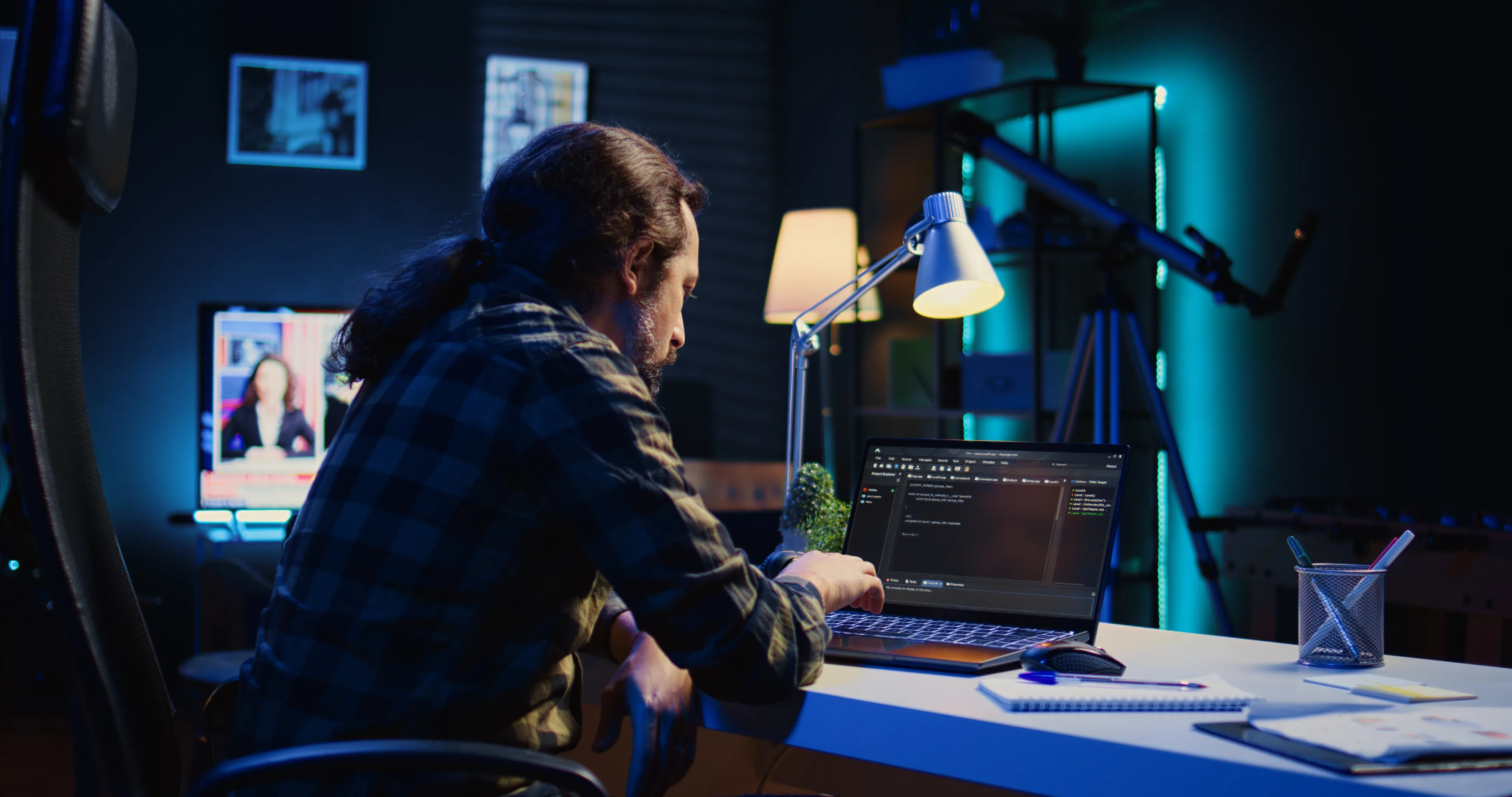 Man in checkered shirt coding on a laptop at his desk under a lamp in a dimly lit room.