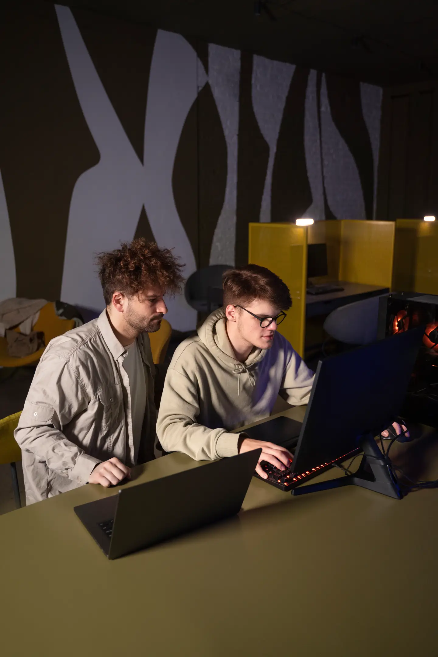 Two men working together at a desk with a laptop and desktop computer in a modern office.
