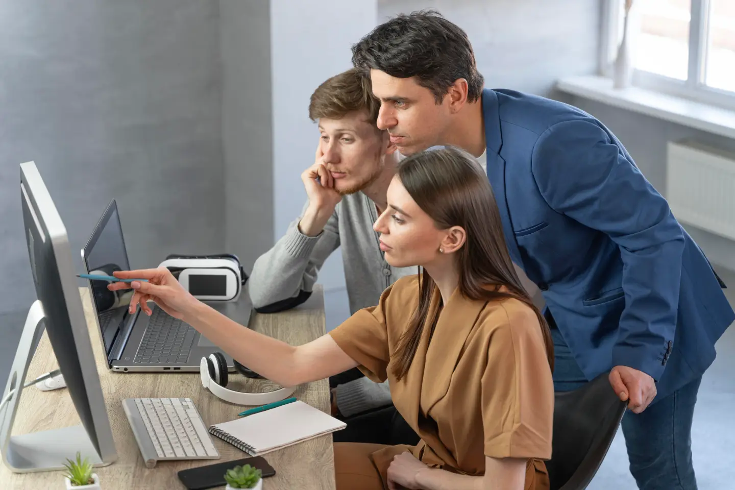 Three colleagues focused on a desktop screen, with one woman pointing at it in a modern office.