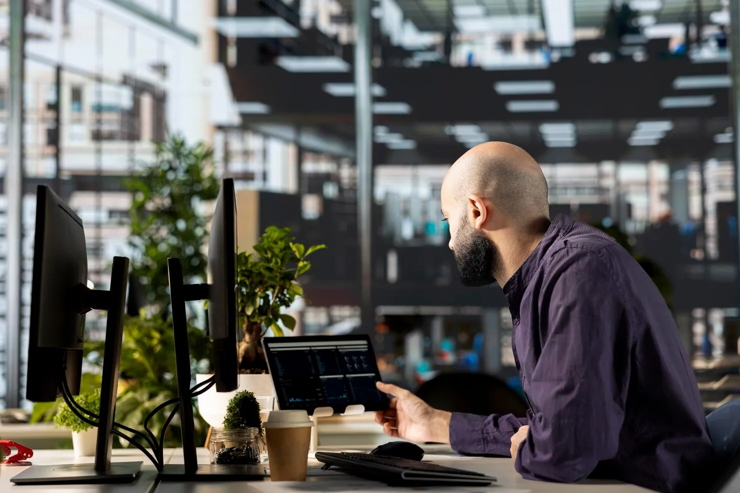 Man with beard working on a tablet at a desk with multiple monitors and plants in a modern office.