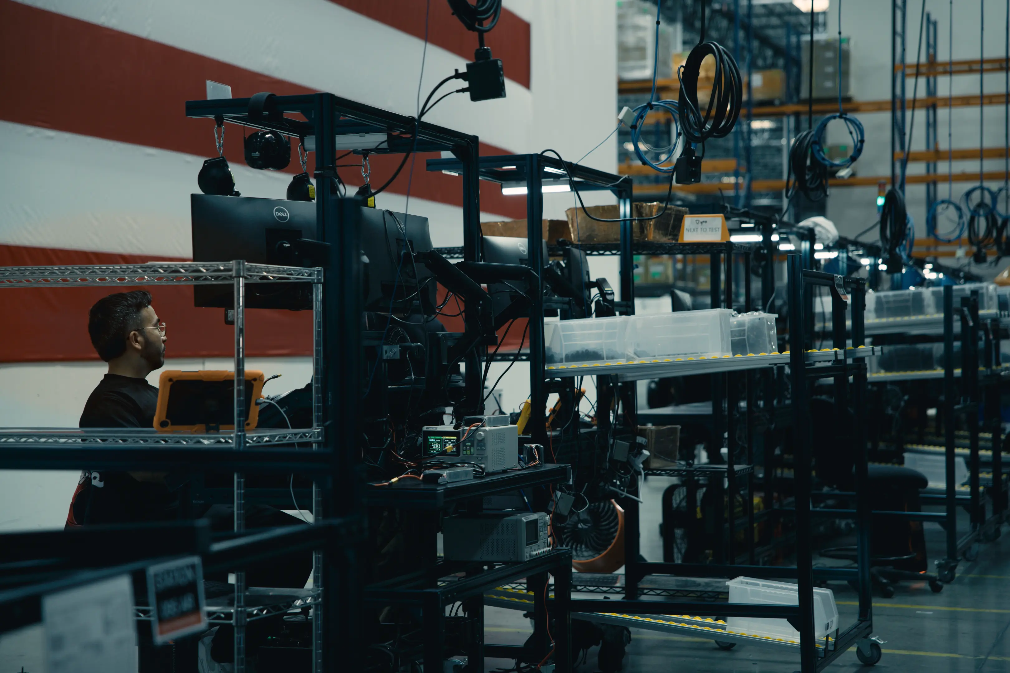 A man sits at a workstation with multiple computer monitors and electronic equipment in an industrial or warehouse setting, surrounded by shelves and hanging cables.