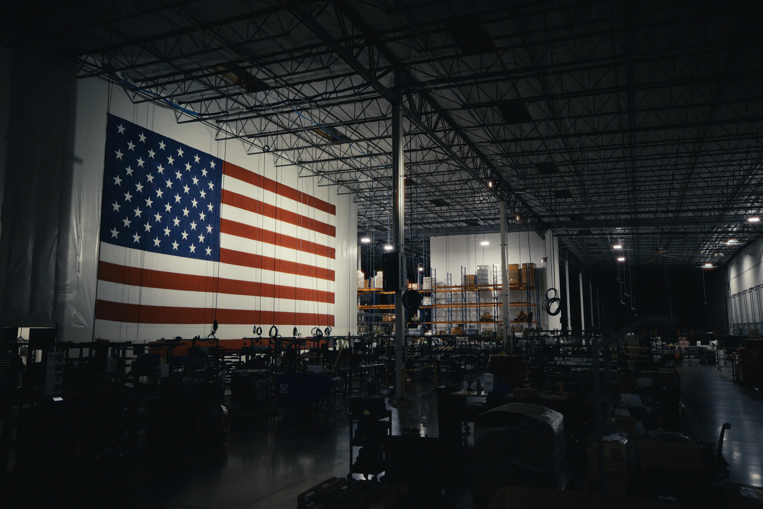 Dimly lit large warehouse interior with a giant American flag hanging on one wall and shelving units stocked with boxes and equipment.