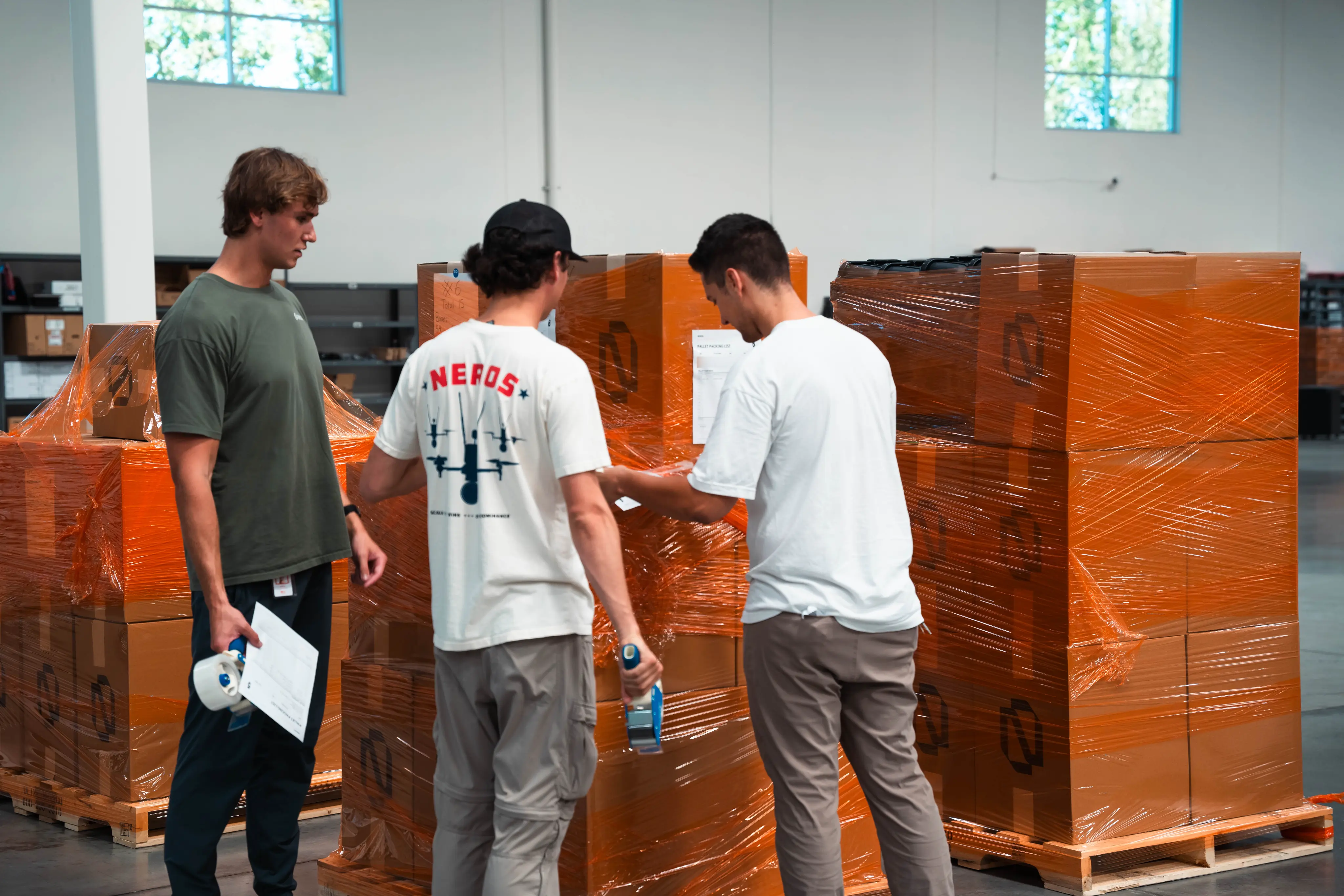 Three men in a warehouse wrapping large stacks of orange boxes secured on wooden pallets with plastic wrap; one man holds packing tape and another holds a sheet of paper, while they appear to be discussing the packaging process.