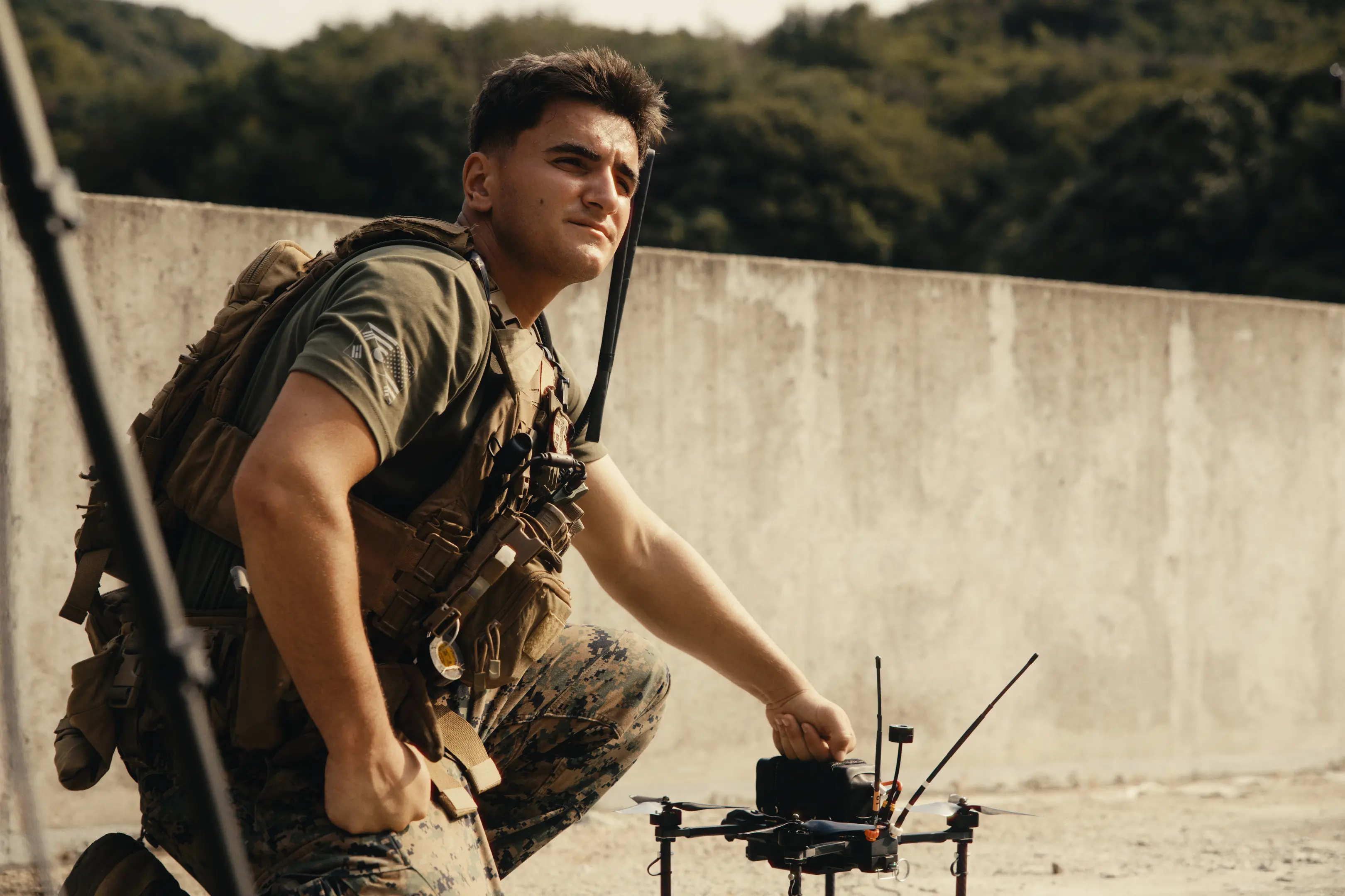 A soldier in camouflage pants and tactical vest crouches outdoors beside a small drone, holding part of its frame, with a concrete wall and forested background.