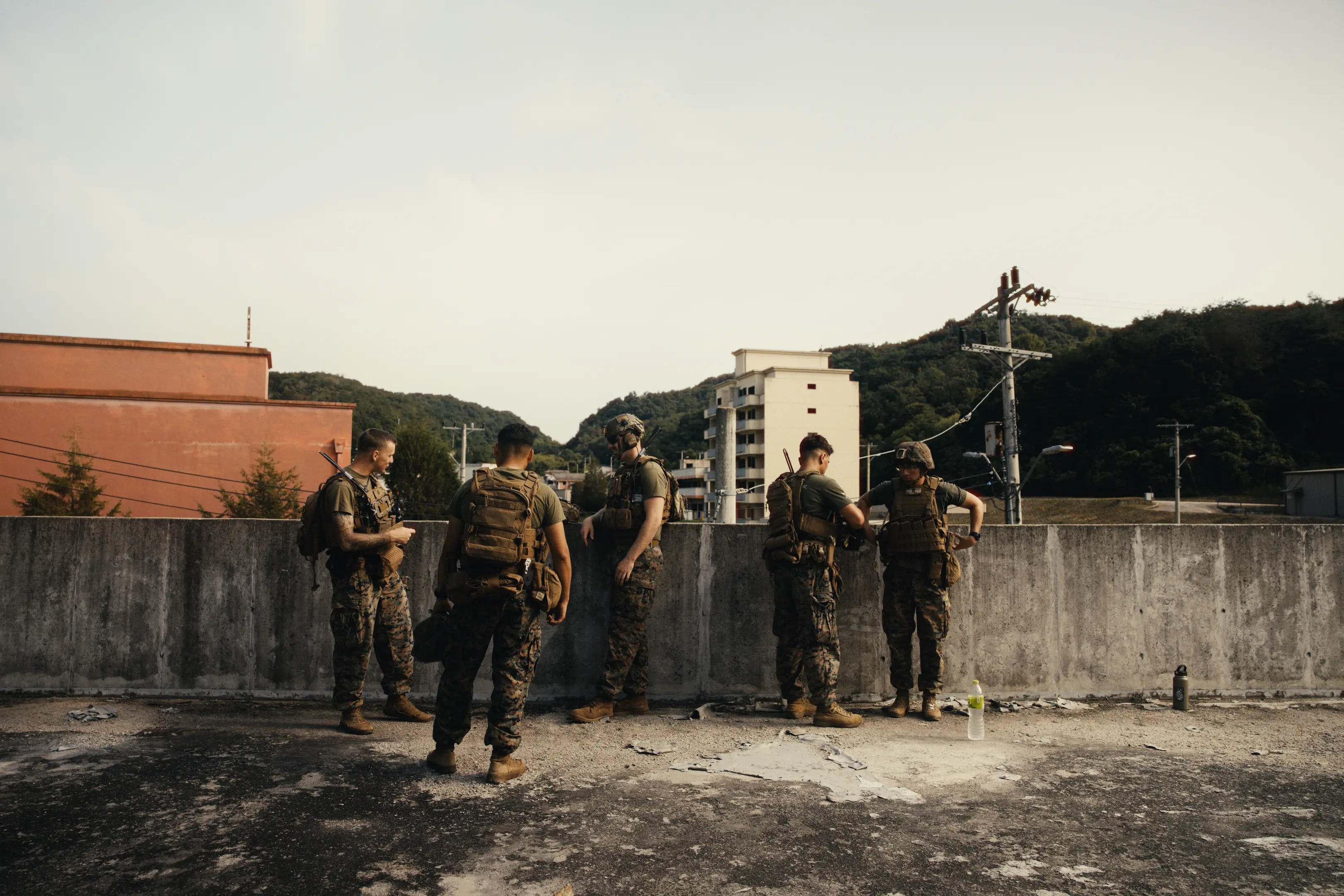 Five soldiers in camouflage uniforms and tactical gear standing and talking on a rooftop with a concrete barrier, with green hills and buildings in the background.
