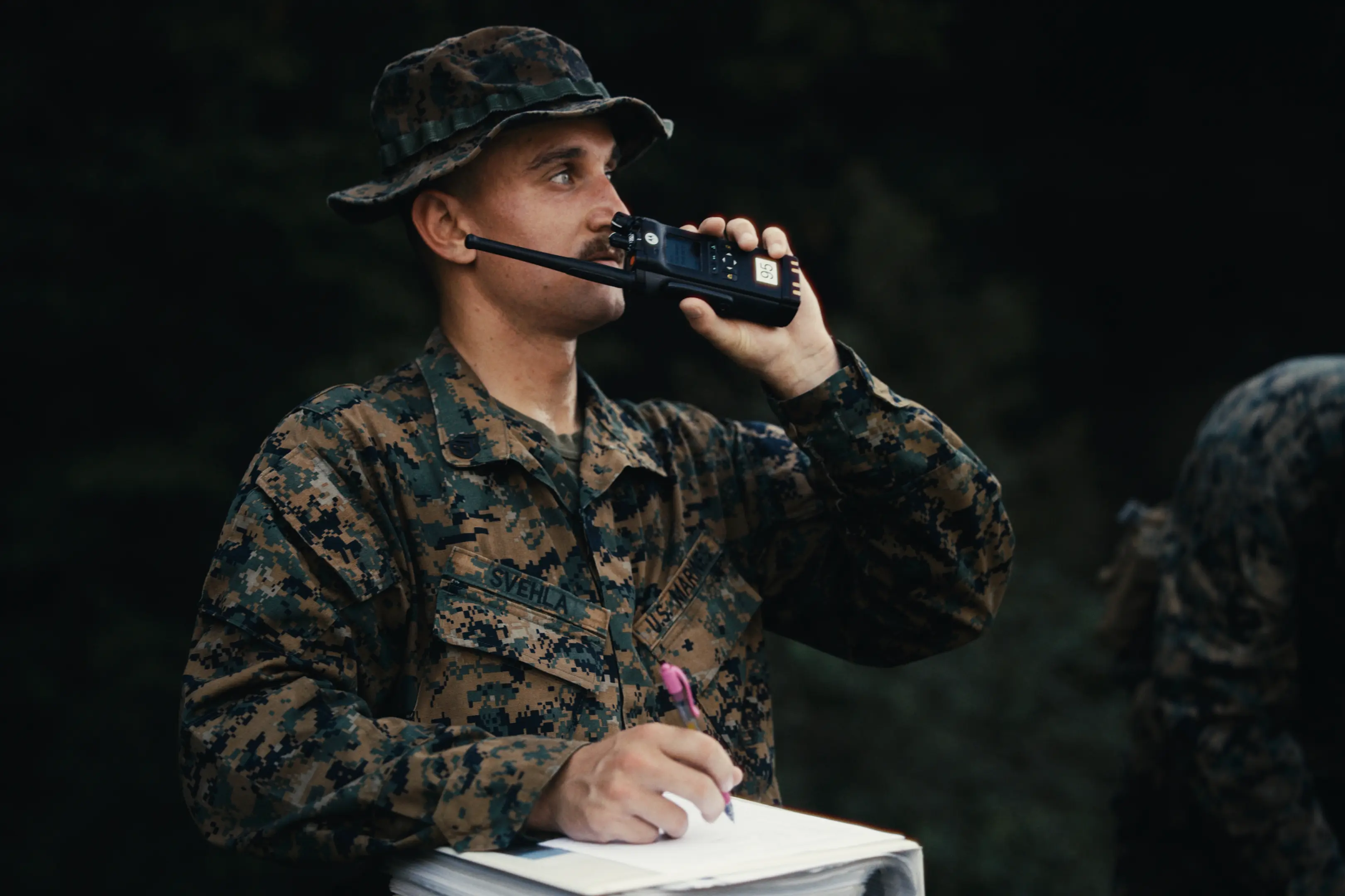 A soldier in camouflage uniform and hat uses a walkie-talkie while holding a pen and writing on a clipboard outdoors.