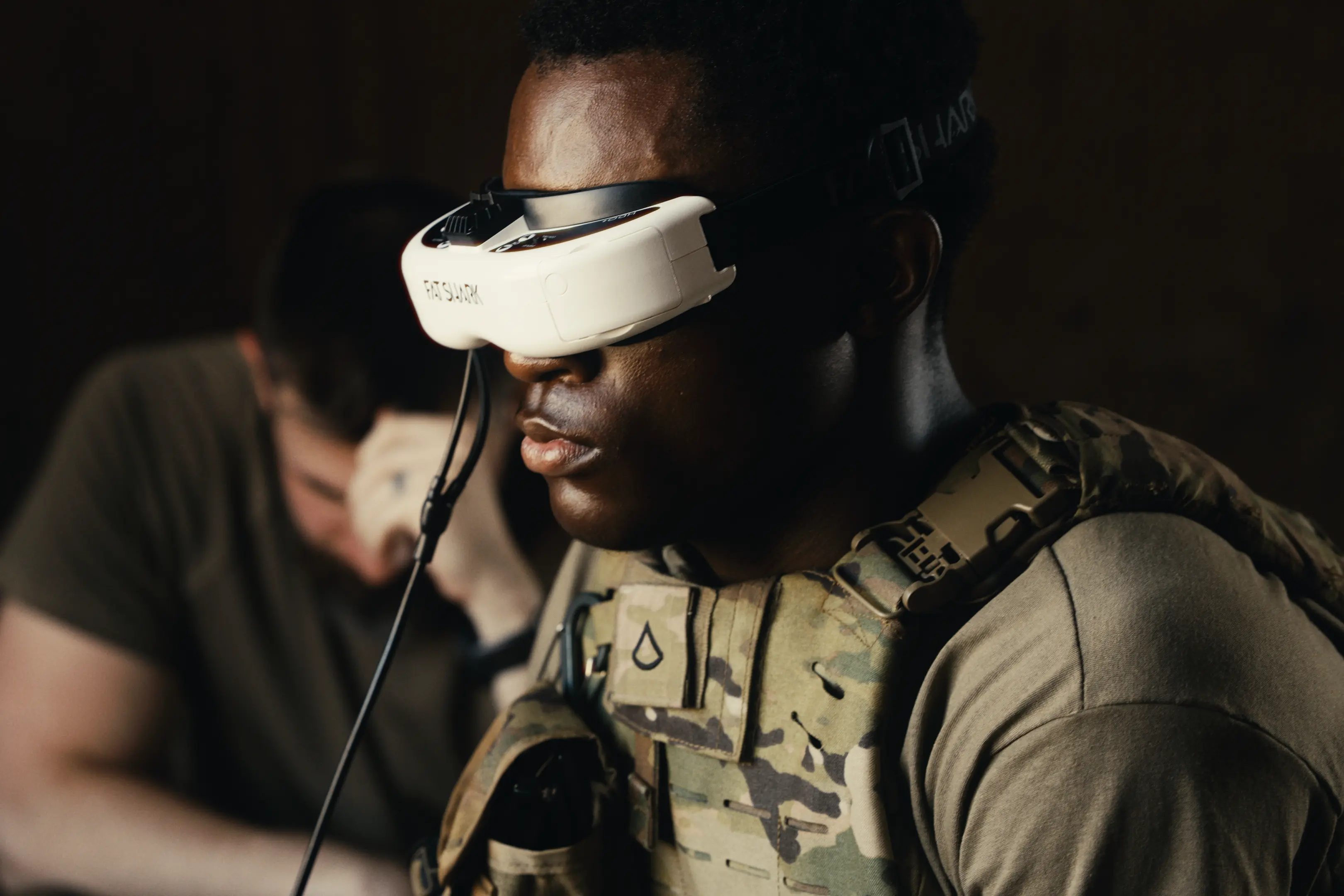 Soldier wearing camouflage tactical gear and a green shirt using white Fat Shark FPV goggles, with another person in the background.