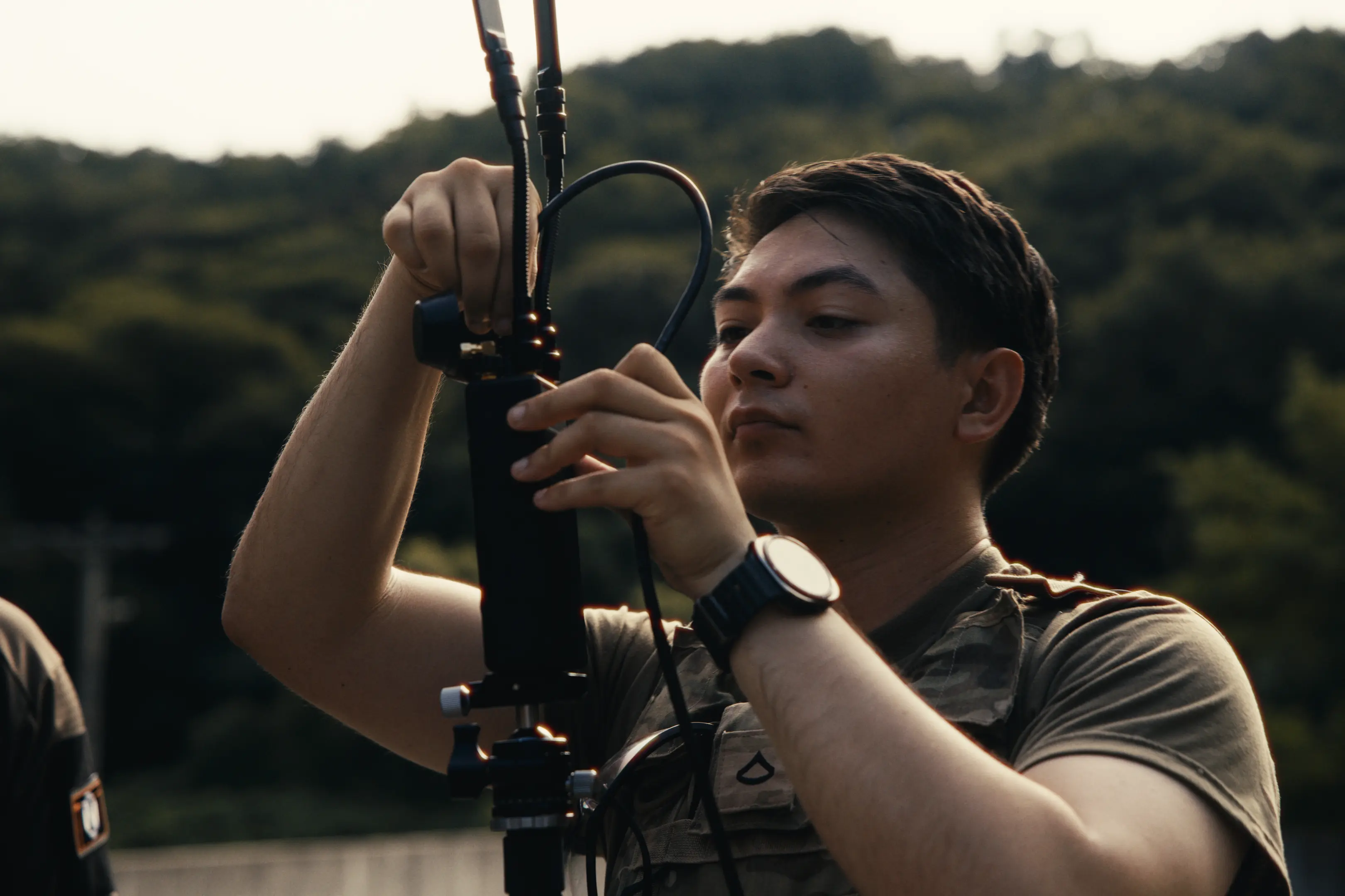Young man outdoors wearing a camouflage vest and watch, adjusting cables on a mounted device.