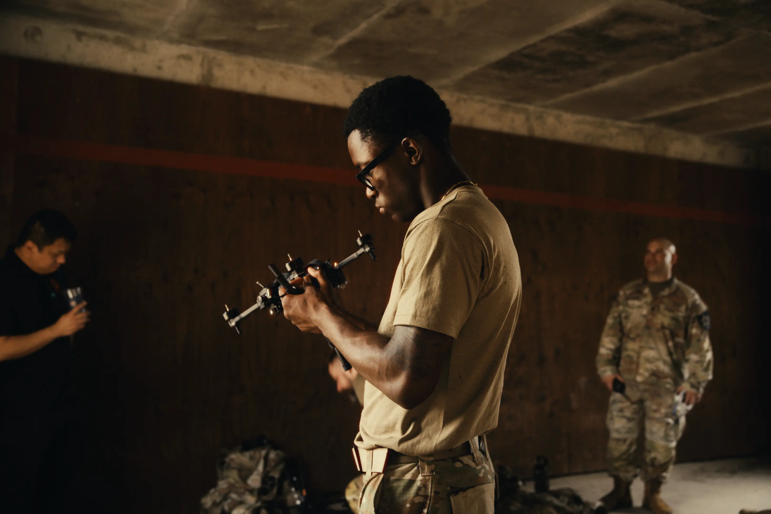 A man in military attire and glasses inspects a piece of equipment indoors, with other people in the background also dressed in military or casual clothing in a dimly lit room.