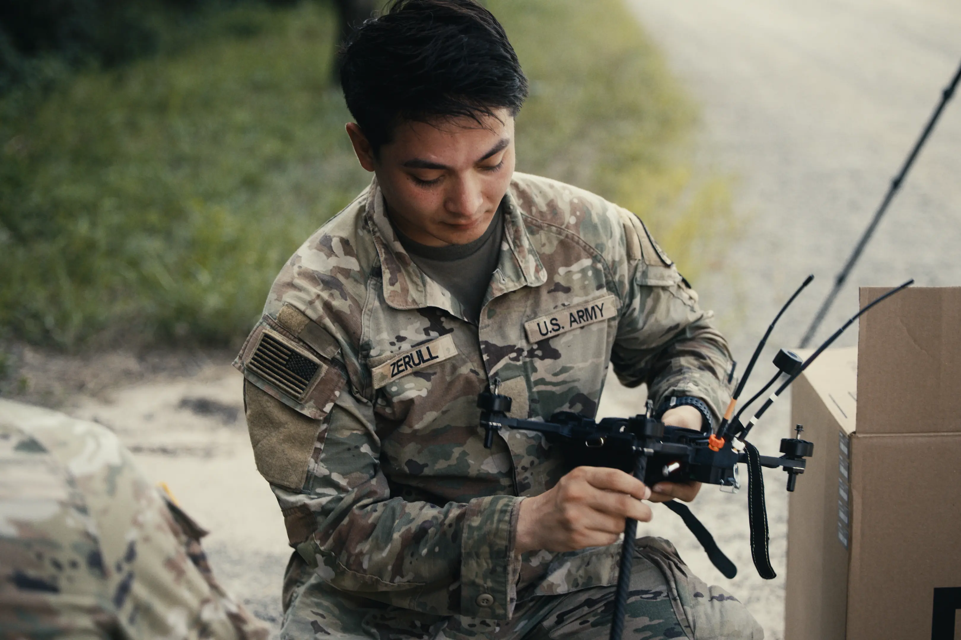 U.S. Army soldier in camouflage uniform kneeling outdoors, handling and examining a small drone with antennae next to a cardboard box.