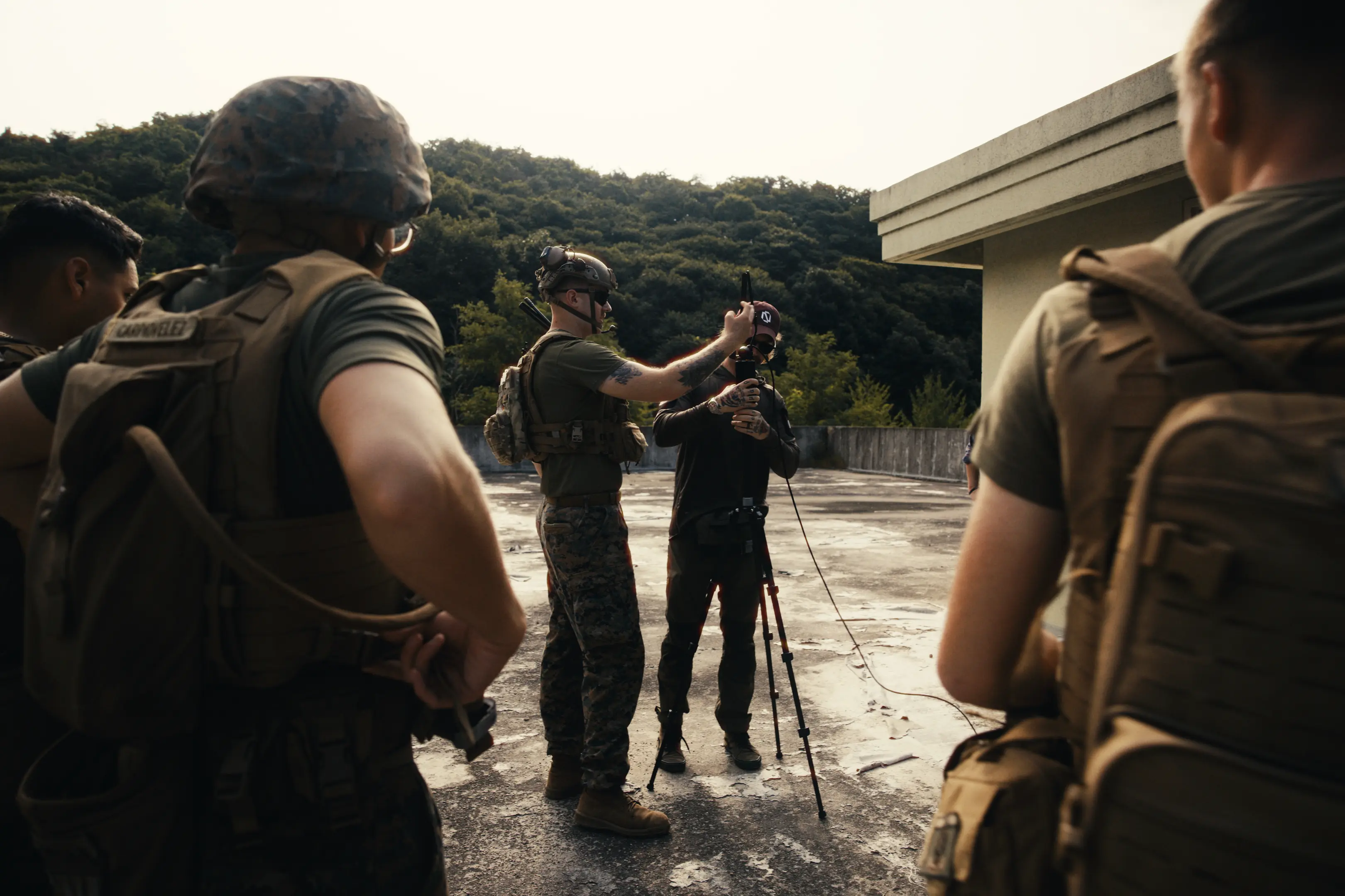 Group of soldiers on a rooftop surrounded by forested hills, with one soldier wearing a helmet adjusting equipment on a tripod while another person in dark clothing assists.