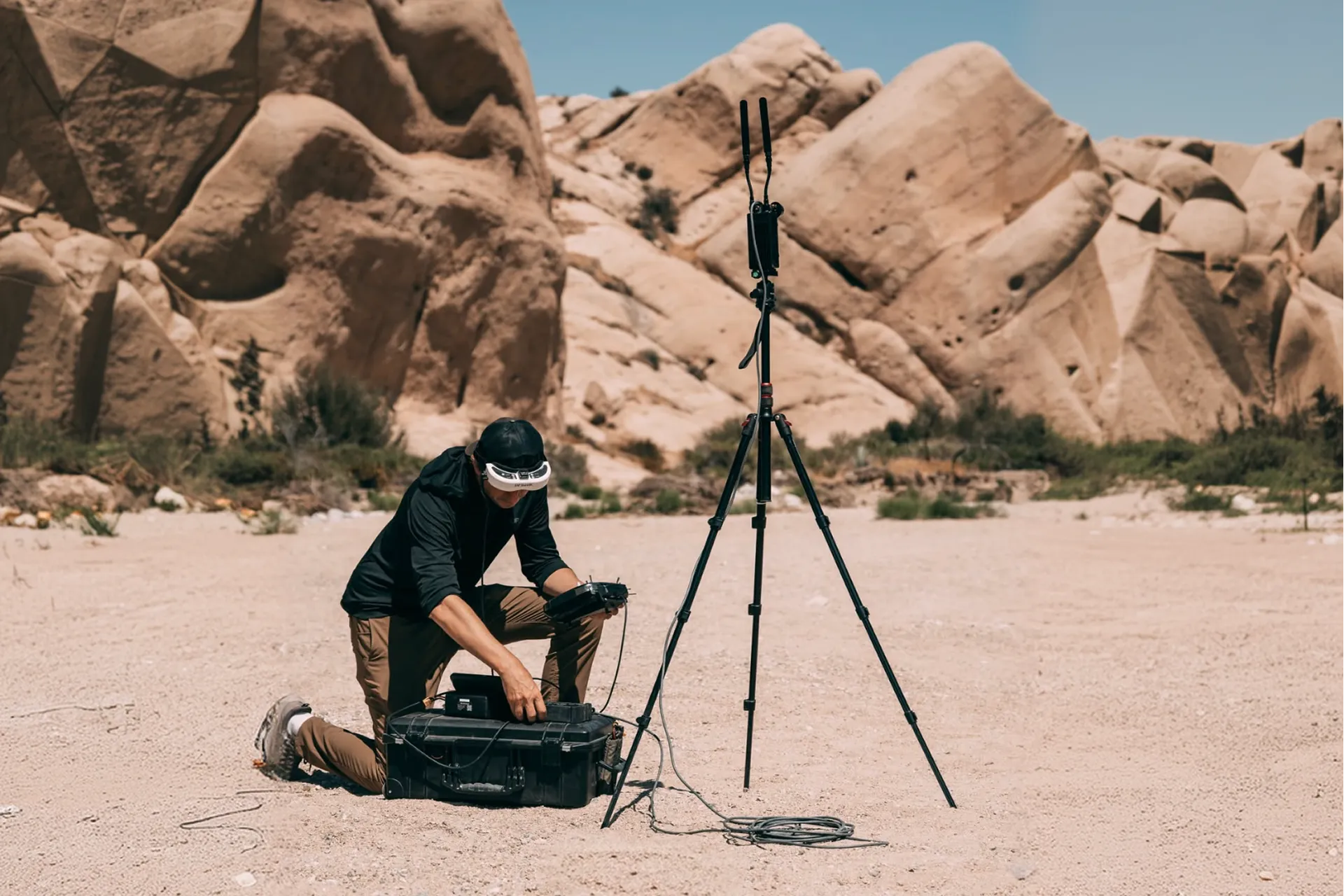 Man kneeling on sandy ground in a rocky desert landscape adjusting equipment inside a black case, next to a tripod with mounted antennas and cables.