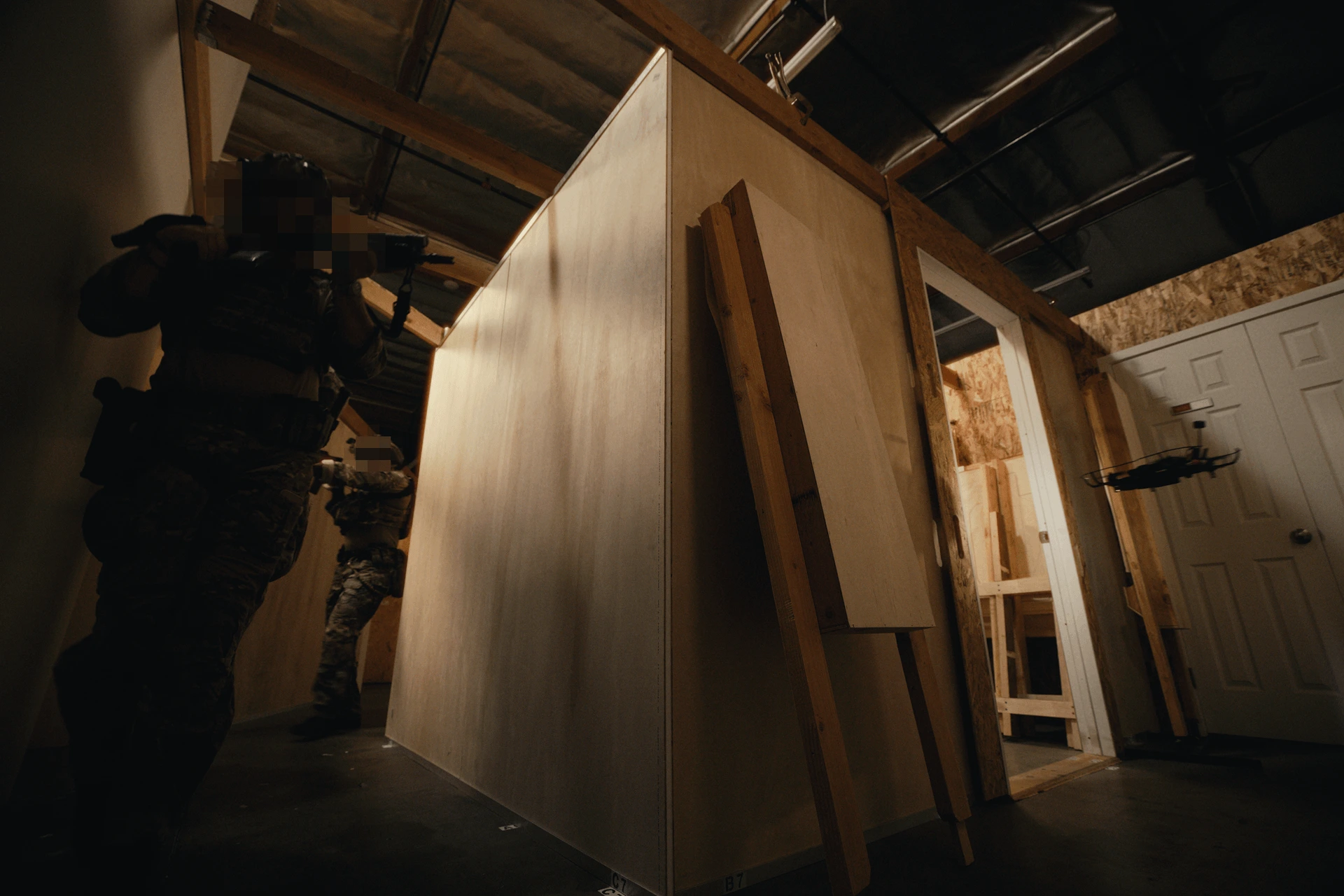 Two armed individuals in tactical gear with blurred faces in a dimly lit training facility with wooden walls; a small drone hovers near an open doorway.