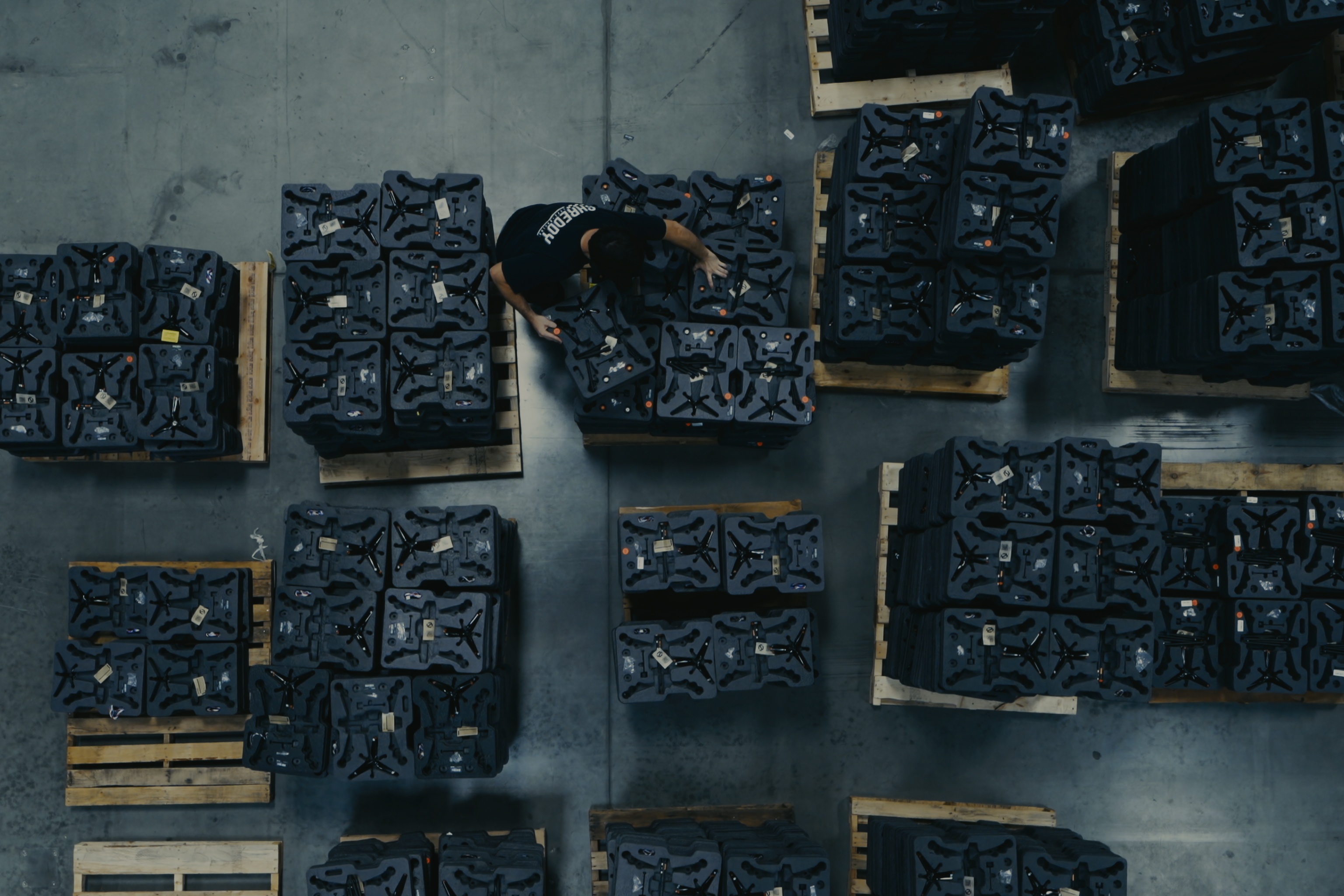 Overhead view of a warehouse worker arranging stacks of black molded trays on wooden pallets inside a large industrial space with a concrete floor.