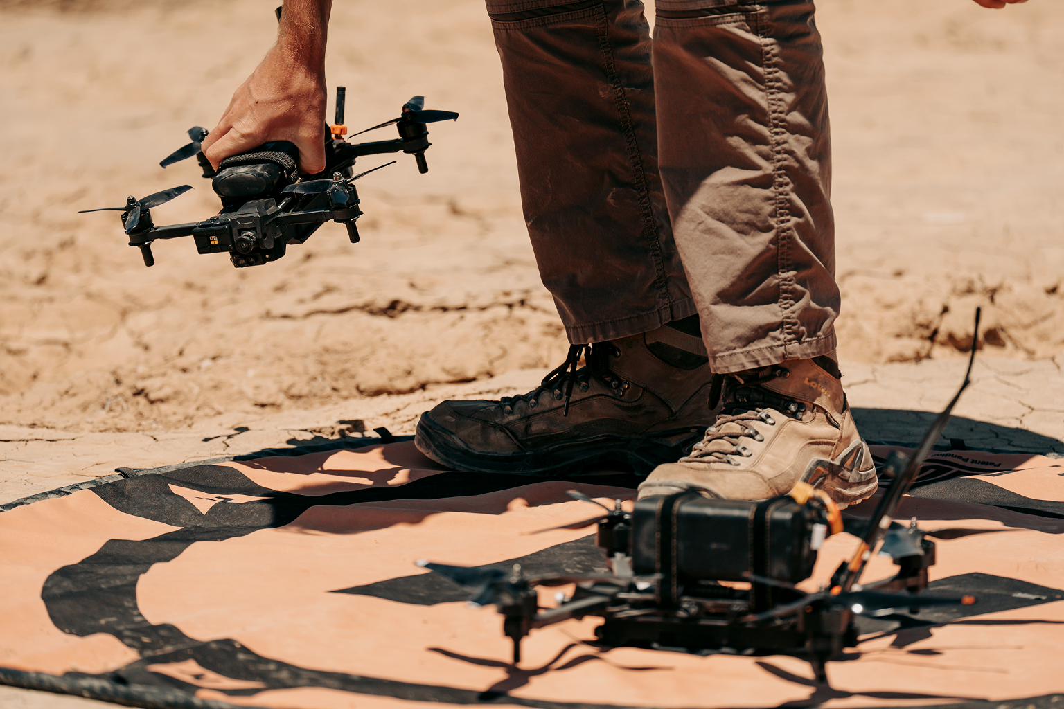 Close-up of a person standing on a mat in a dry, cracked landscape, holding a small black drone in one hand while another drone rests on the mat near their feet.